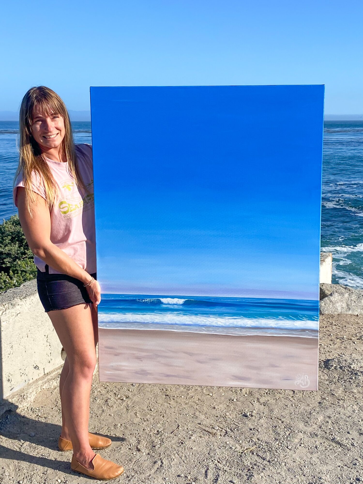 Woman holding a large painting of a beach scene on a sandy shore with ocean and blue sky in background.