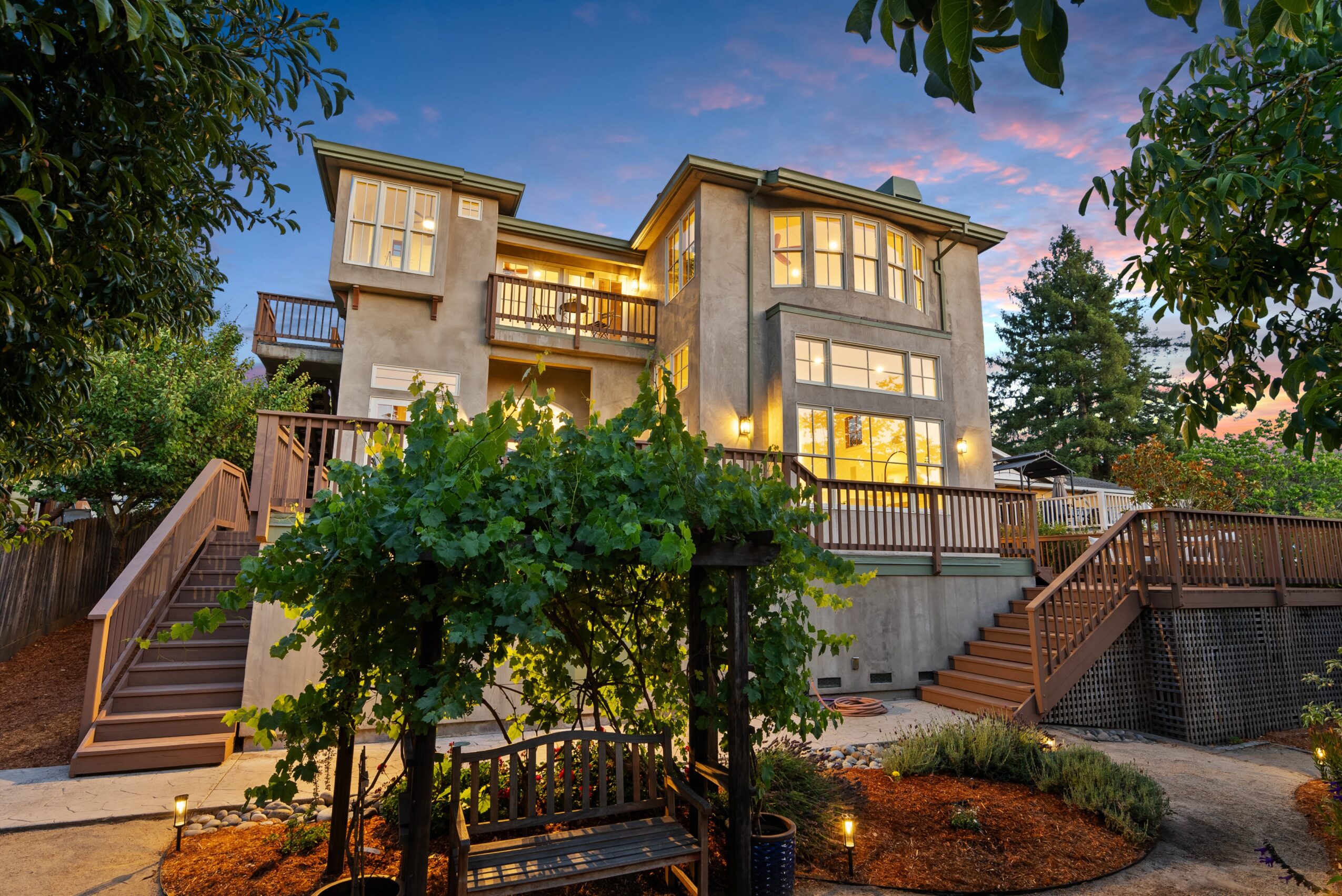 Multi-story house with large windows, surrounded by trees and a landscaped yard, during sunset with illuminated interior lights.