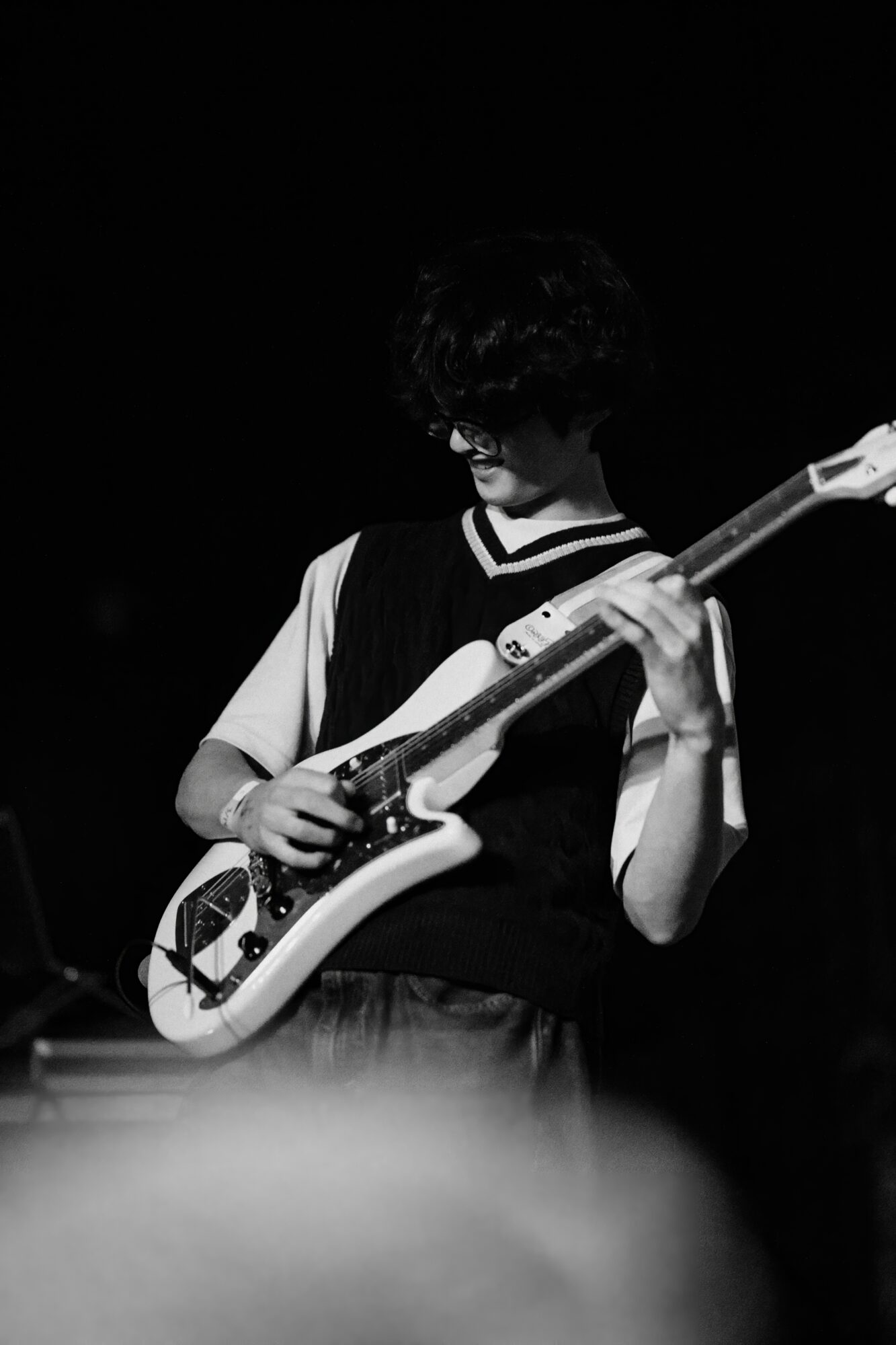 Person playing electric guitar on stage, with curly hair and dark background.