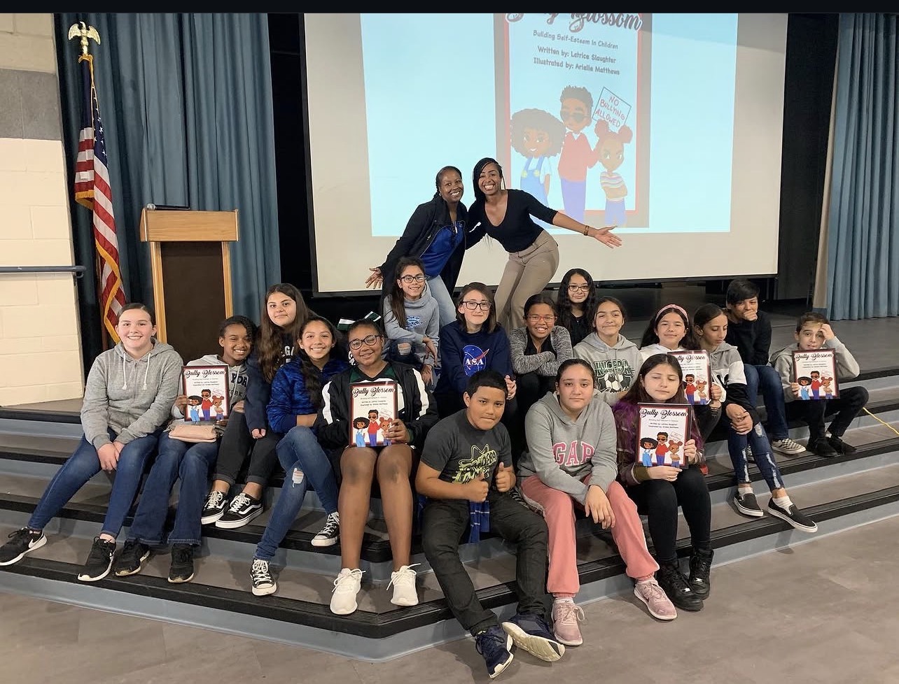 Group of children and two adults on stage, holding books, with a presentation screen and American flag in background.