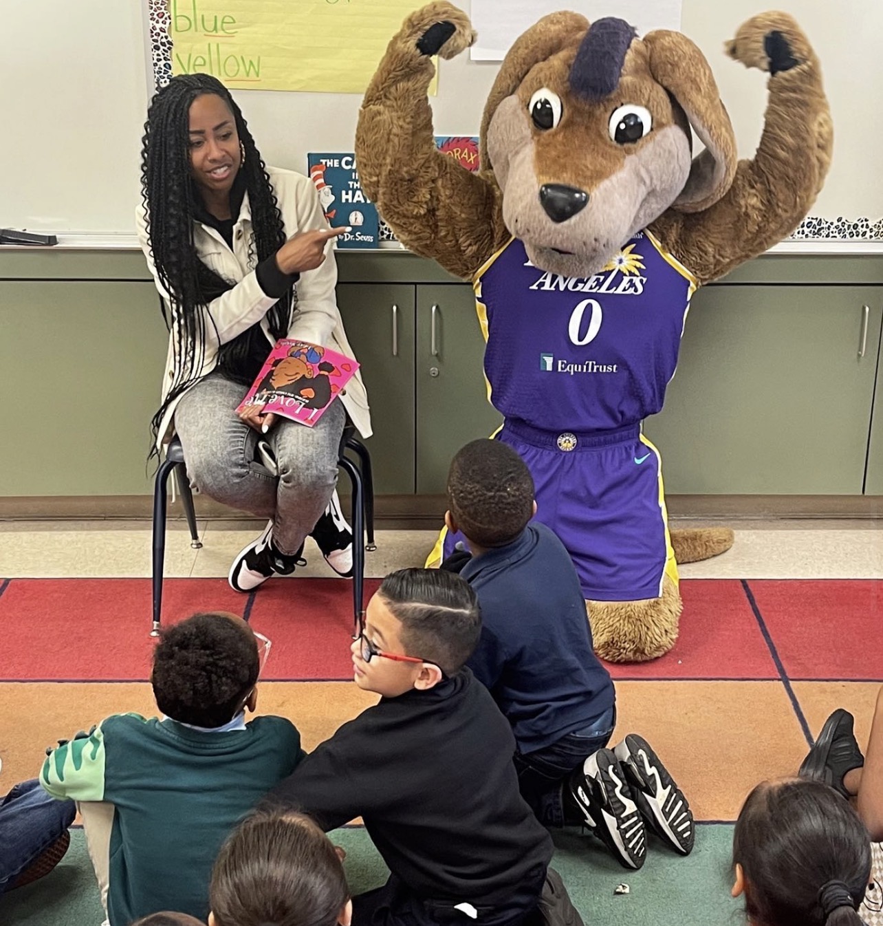 Person sitting on a chair pointing at a mascot with a dog costume, children sitting on the floor watching.