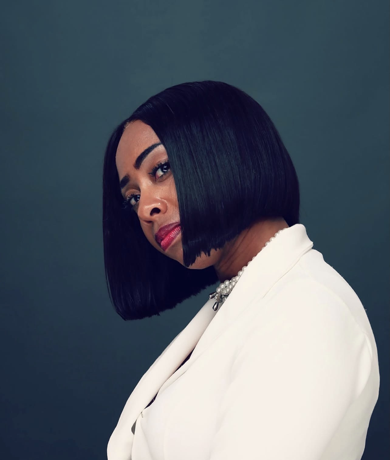 Woman with straight black bob haircut wearing a white blazer, looking sideways against a dark background.
