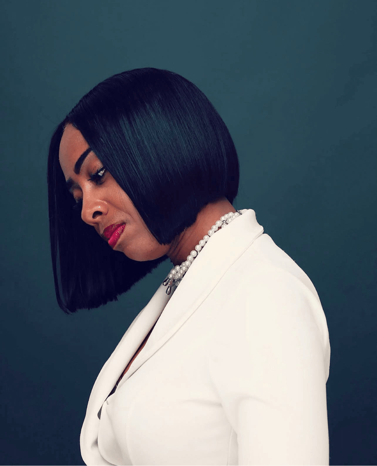 Woman with straight black hair in a bob, wearing a white blazer and pearl necklace, looking down against a dark background.