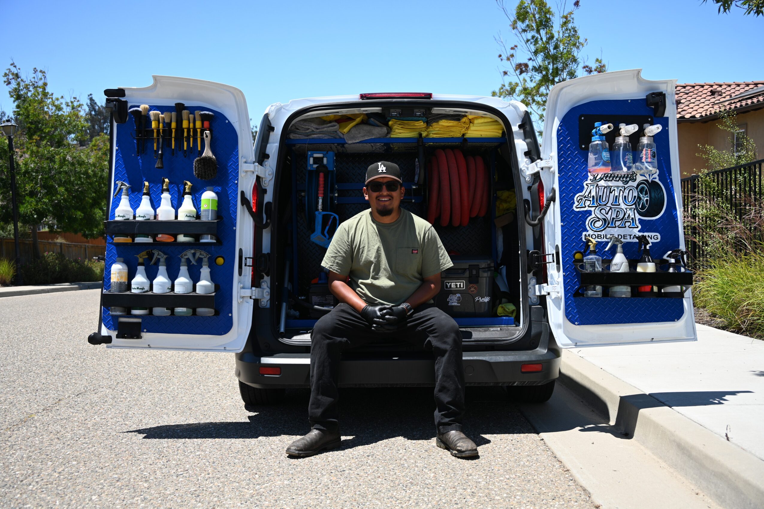 Man sitting in open van with tools and equipment, doors open, outdoor setting, sunny day.