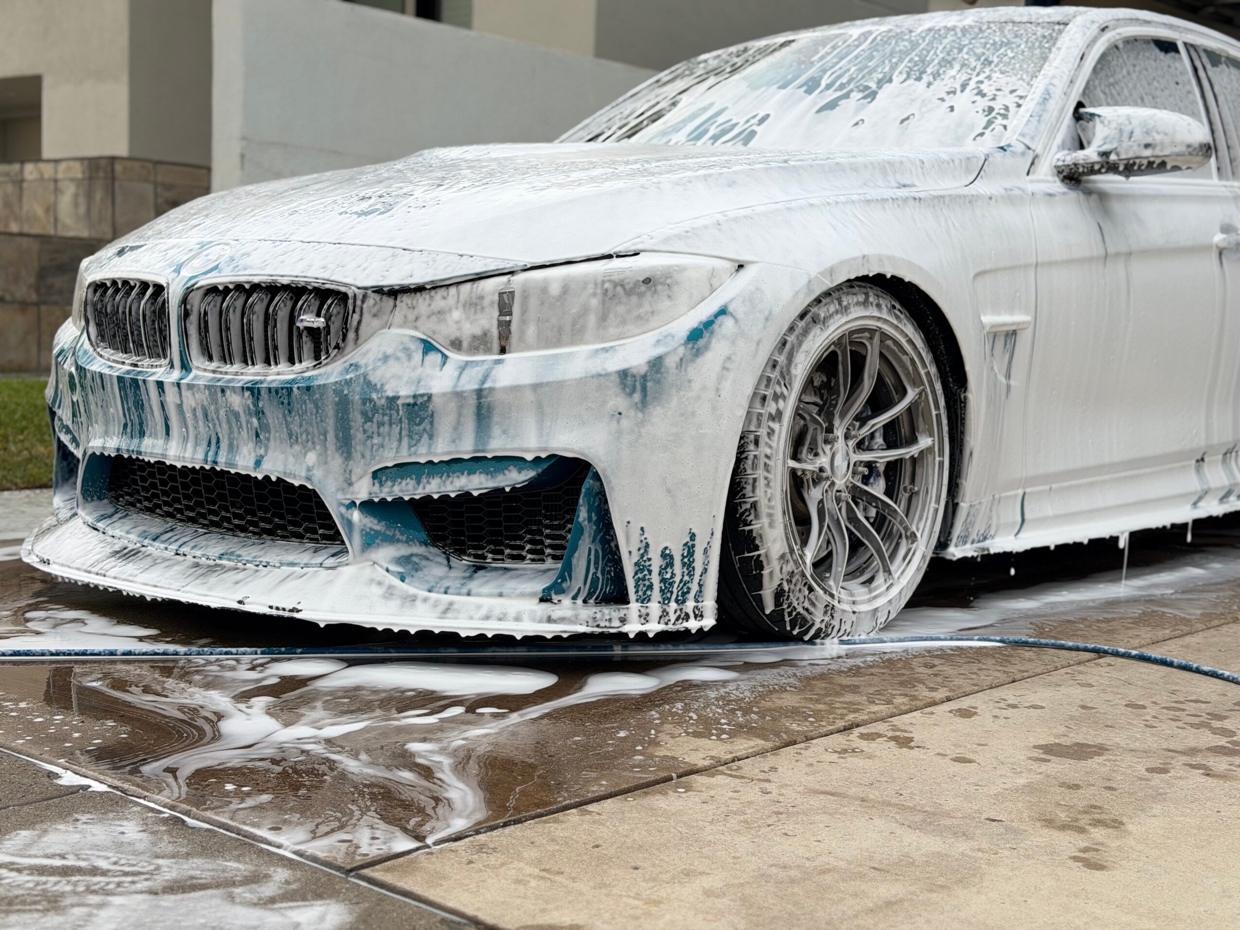 Car covered in soap suds during washing, with a wet driveway and building in background.
