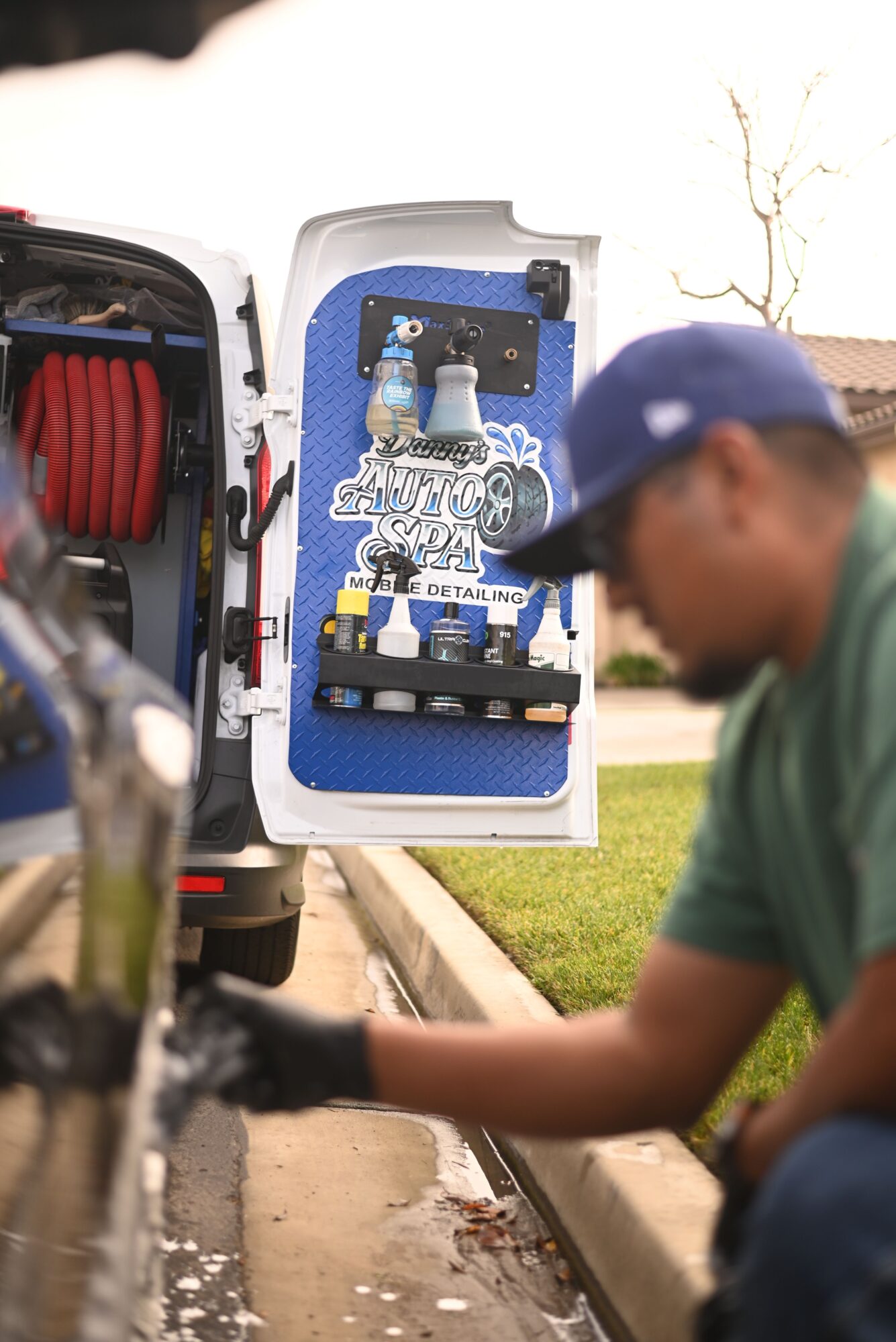 Person in green shirt and blue cap working with a tool kit outdoors, with a grassy area and sidewalk nearby.