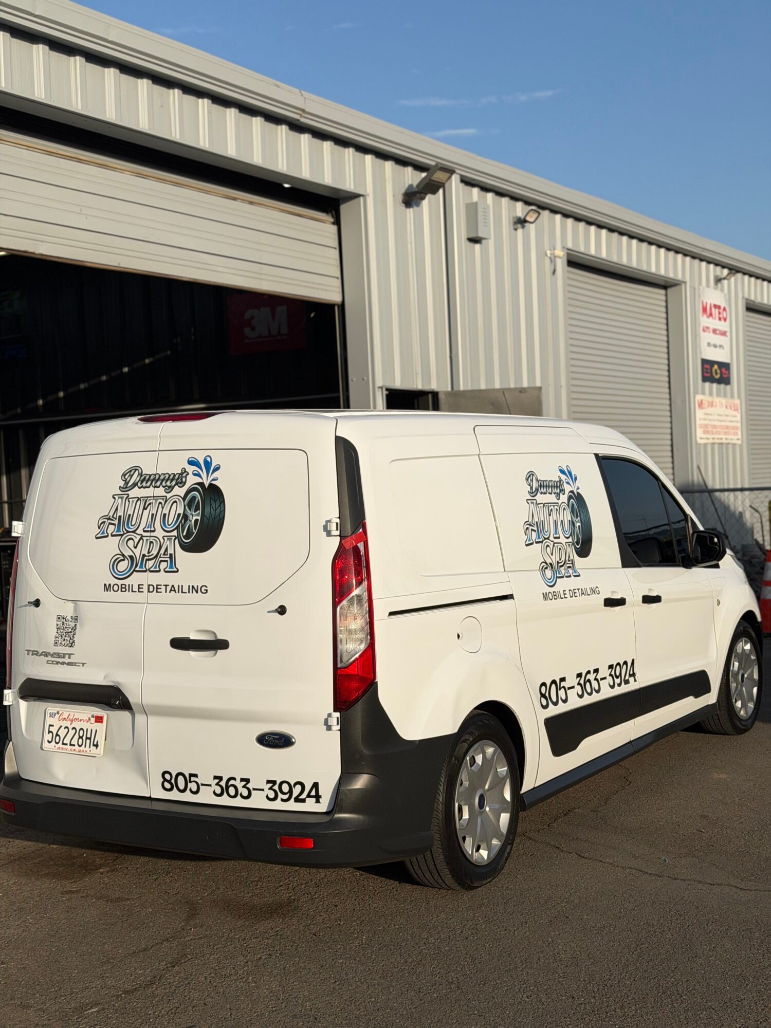 White van parked outside a metal building with two closed garage doors, logo and contact info on the side, clear sky.