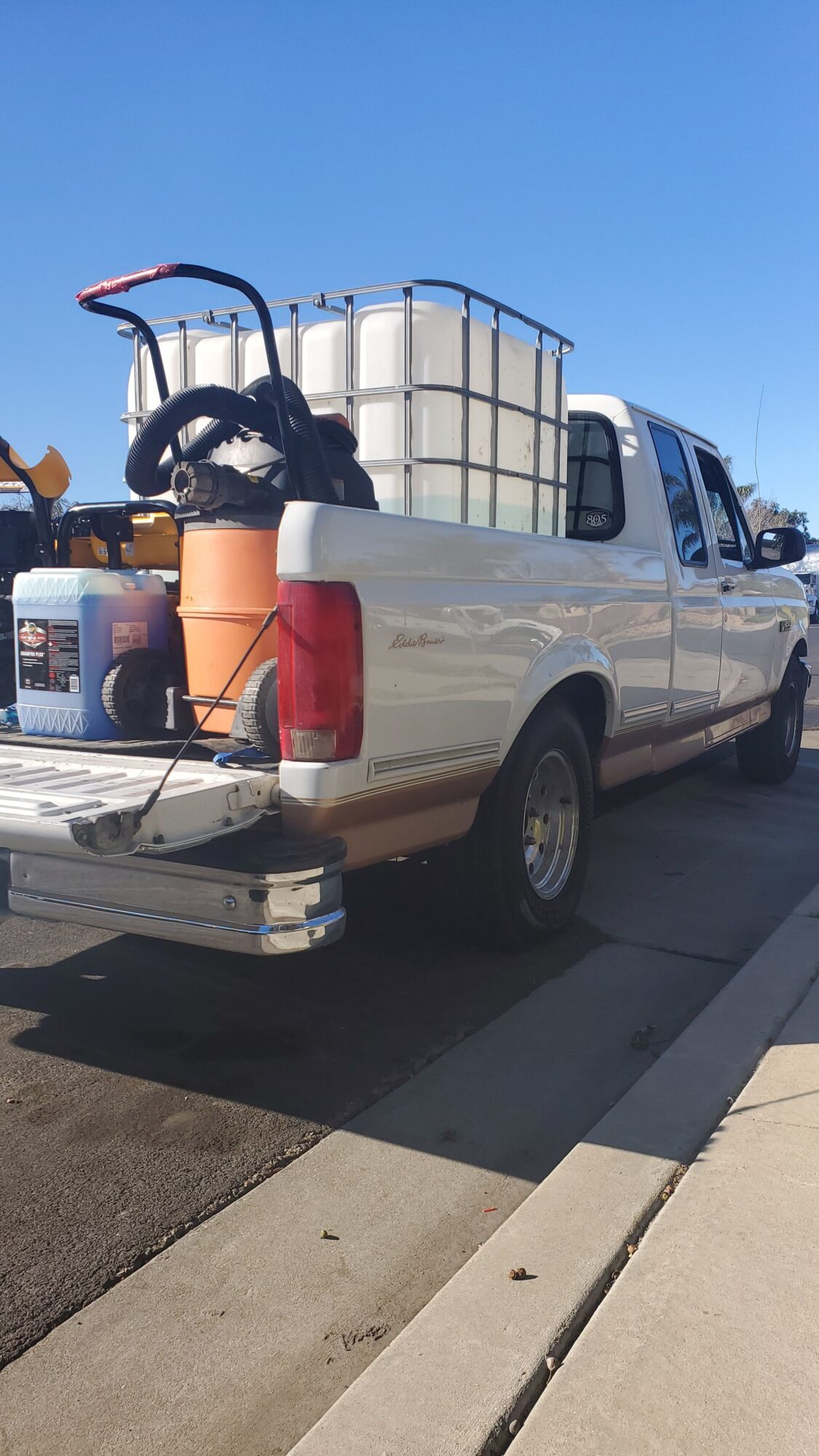 Pickup truck with equipment and supplies parked on a sidewalk, shadow cast on ground, clear blue sky.