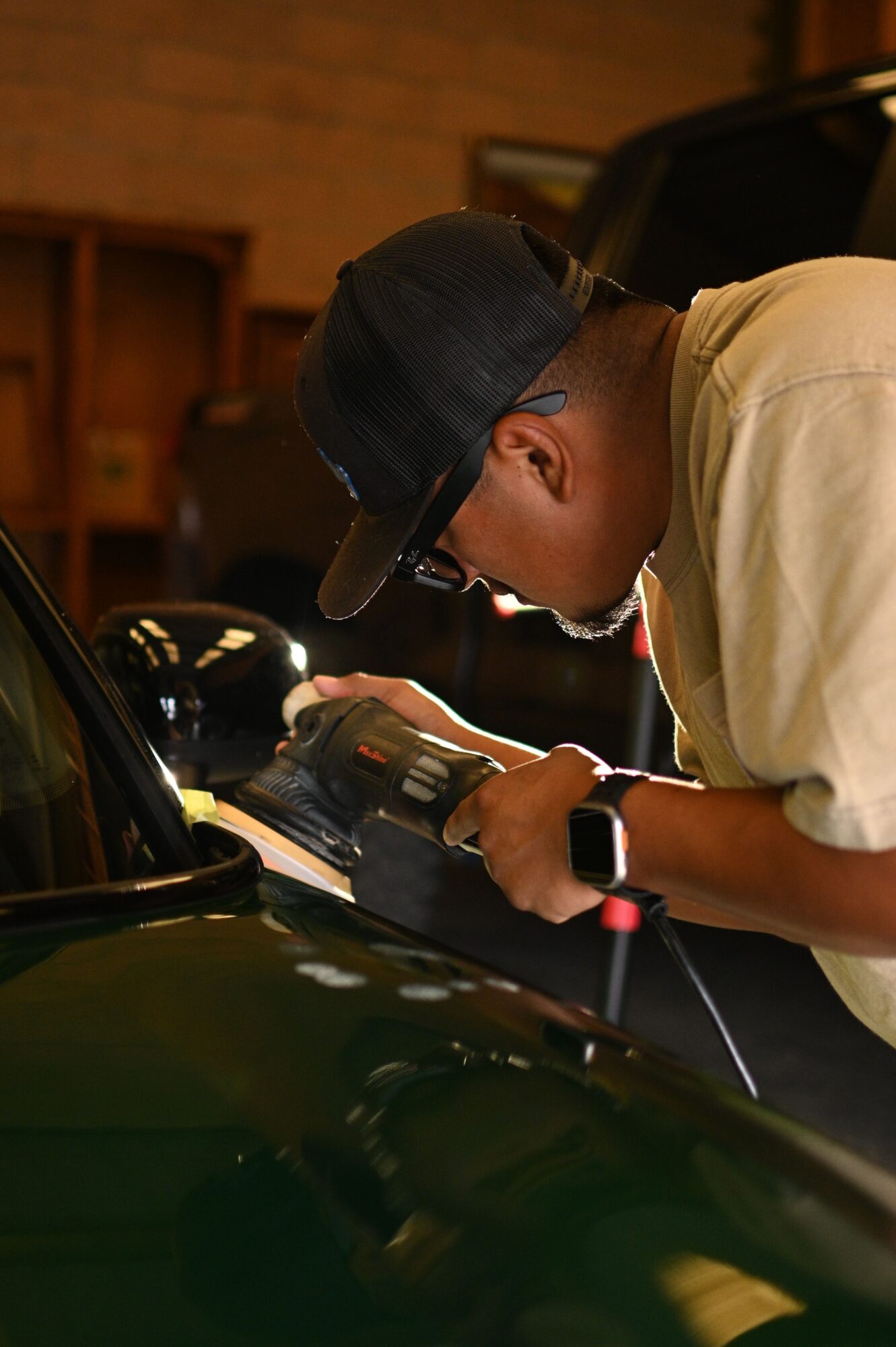 Person wearing a cap and glasses, using a camera or device, sitting in a vehicle interior, with a wooden background.
