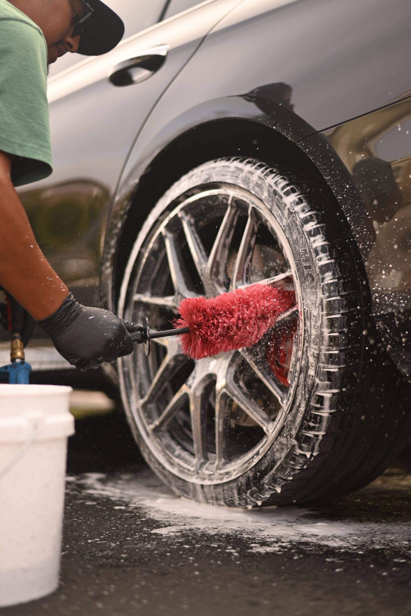 Person cleaning car wheel with red brush, water splashing, wearing black glove, green shirt, and cap.