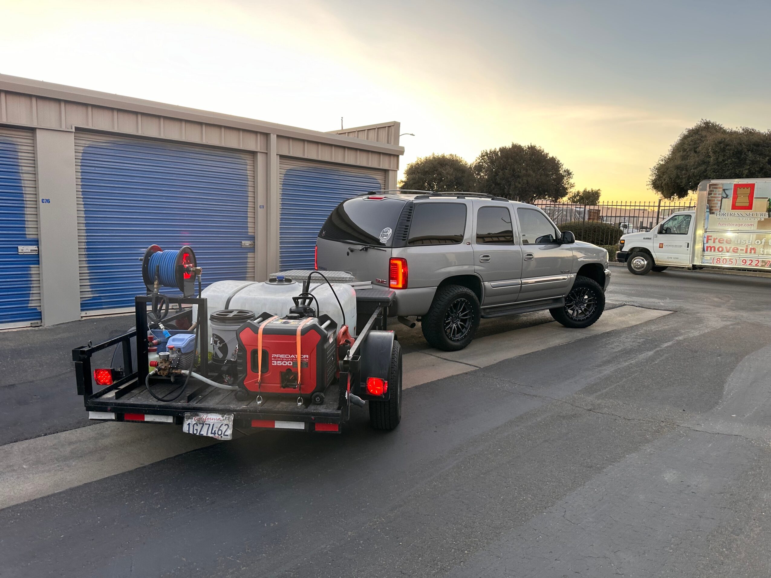 Silver SUV parked in front of storage units, trailer with equipment attached, and other vehicles in the background.