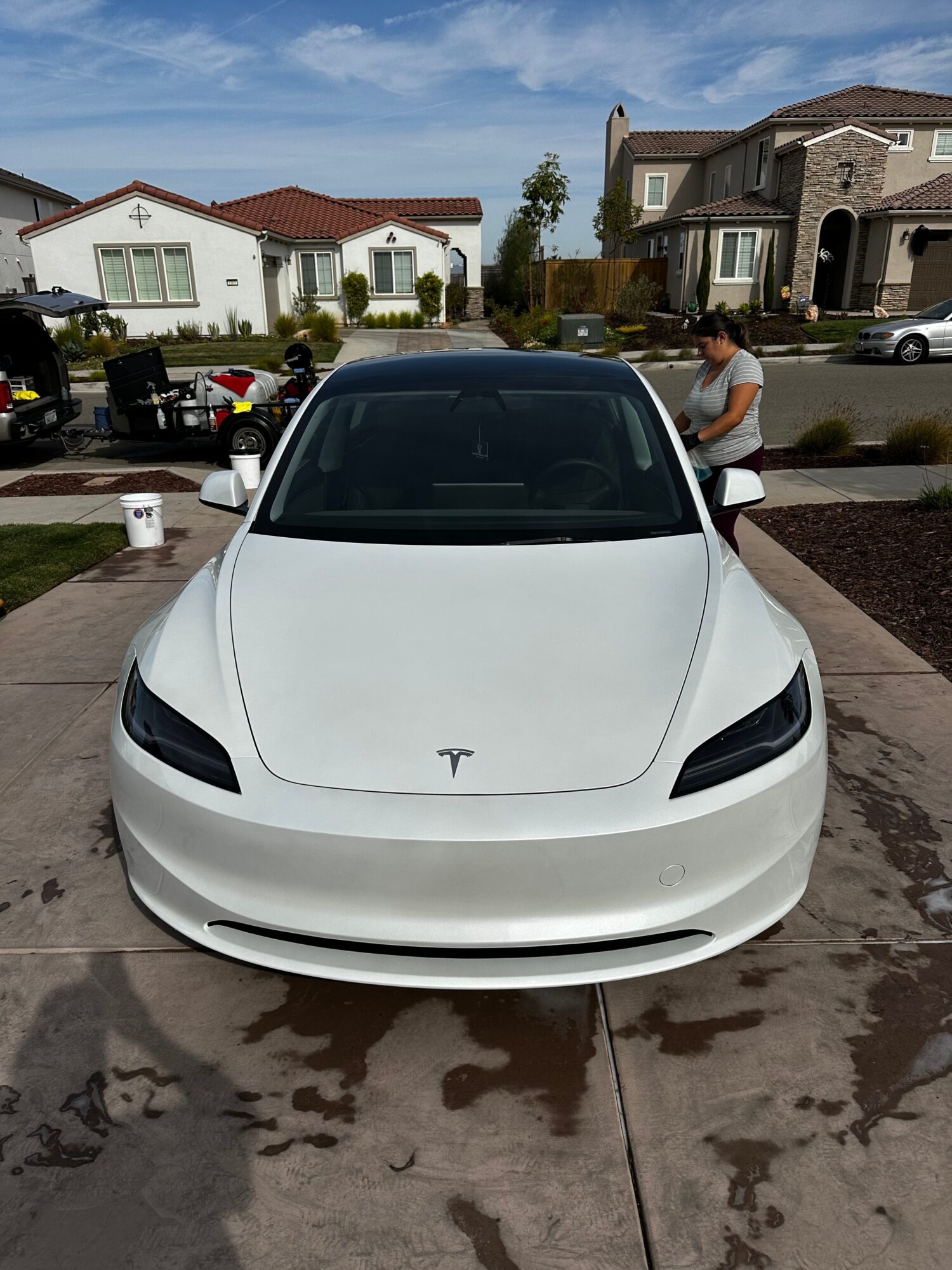 White Tesla car parked on driveway with house and trees in background.