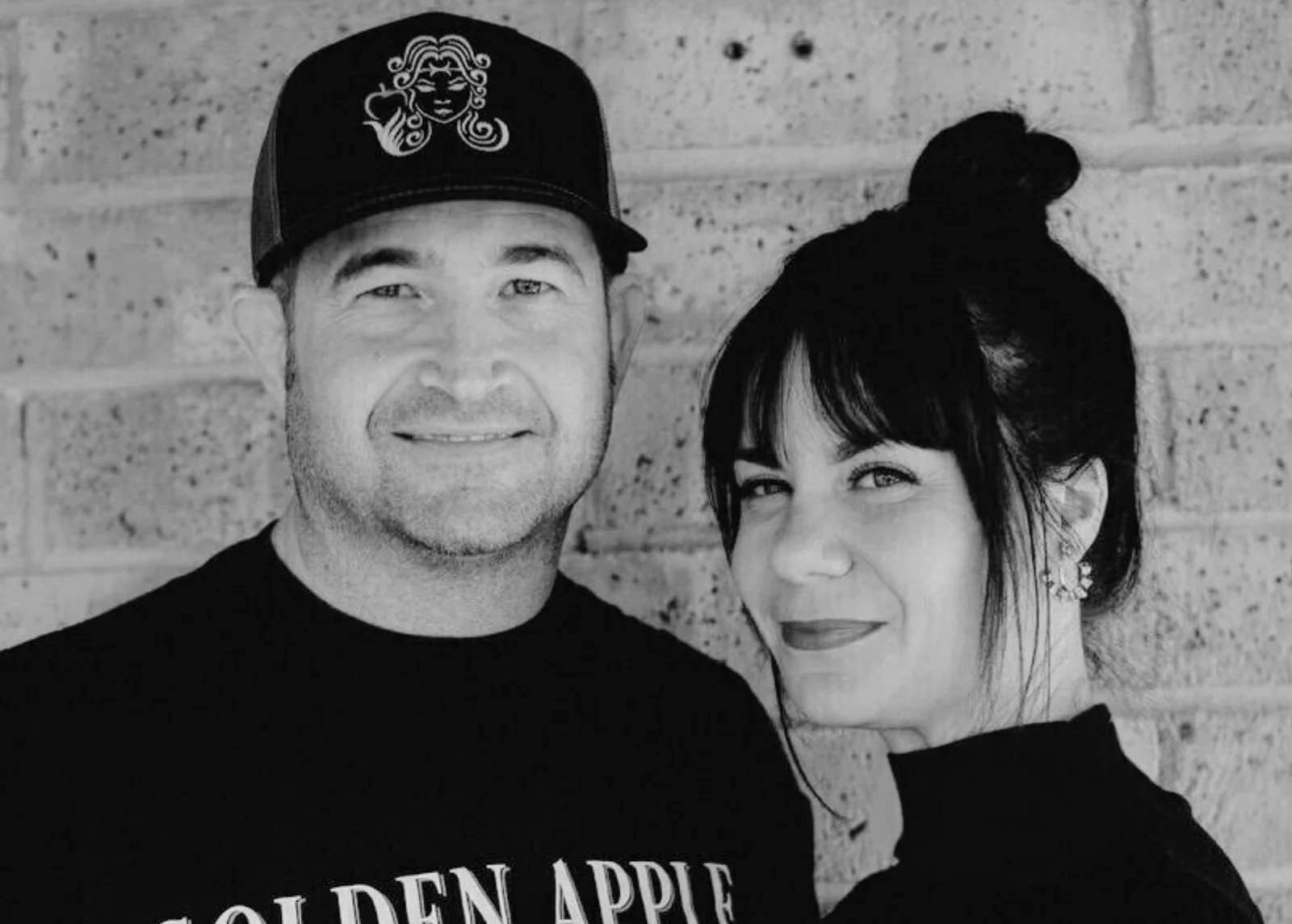 Two people smiling, one wearing a cap and the other with hair in a bun, standing against a brick wall.