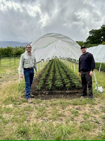 Two people stand in front of a greenhouse with plants inside, outdoors on a cloudy day, with trees and mountains in background.