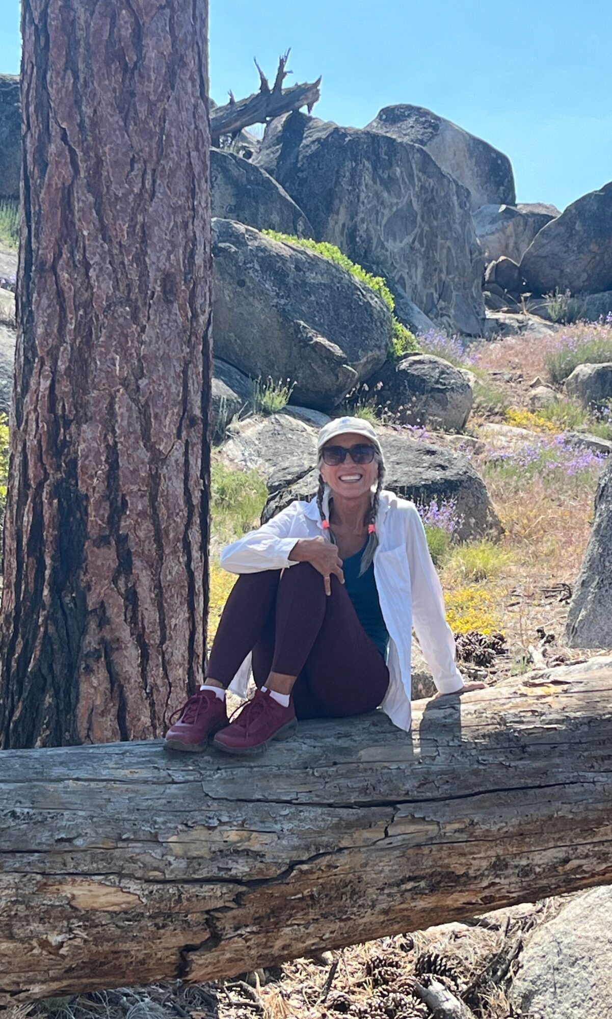 Person sitting on a fallen log next to a large tree in a rocky outdoor area, smiling with sunglasses, blue sky above.
