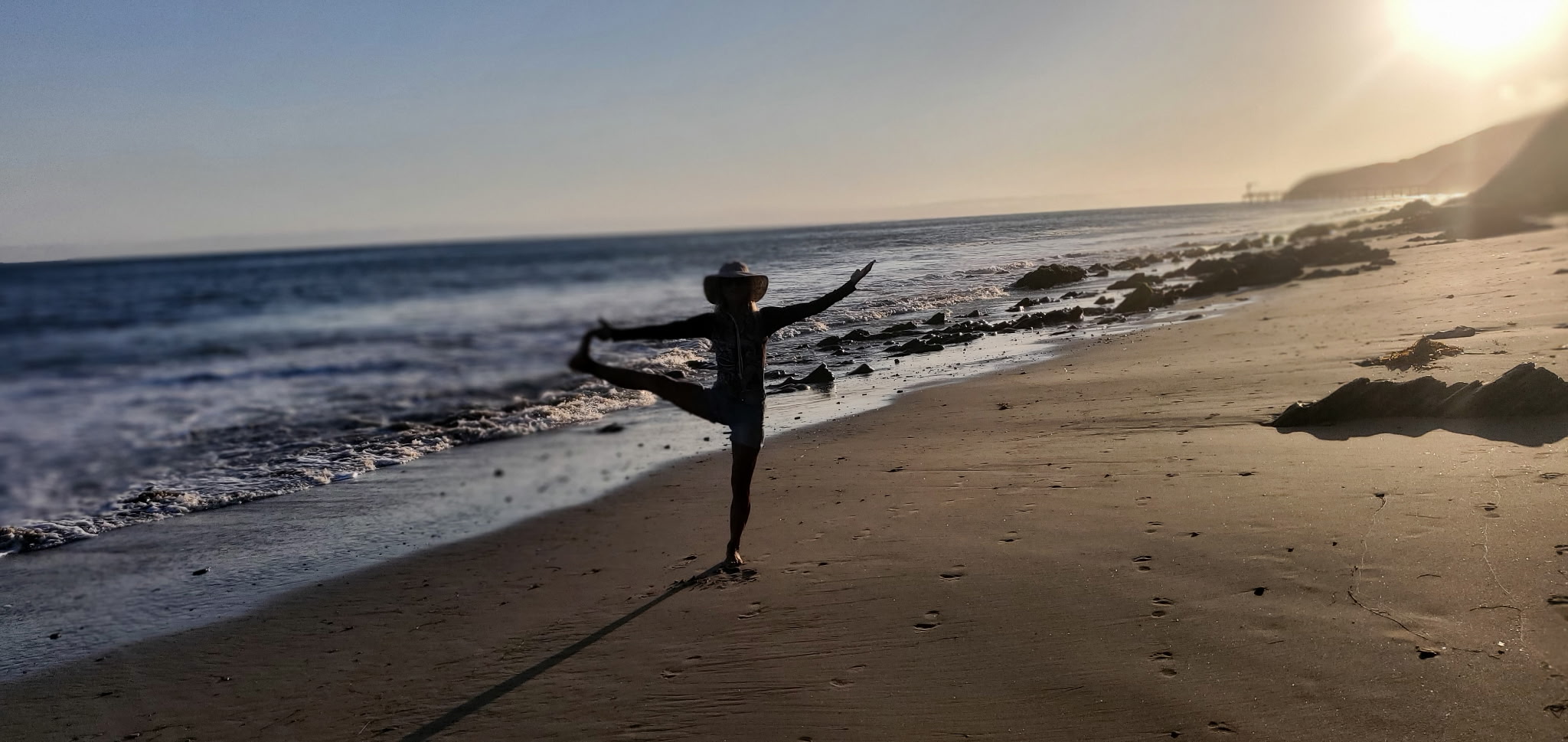 Person standing on beach with arms outstretched, facing the ocean during sunset, with waves and rocky shoreline visible.