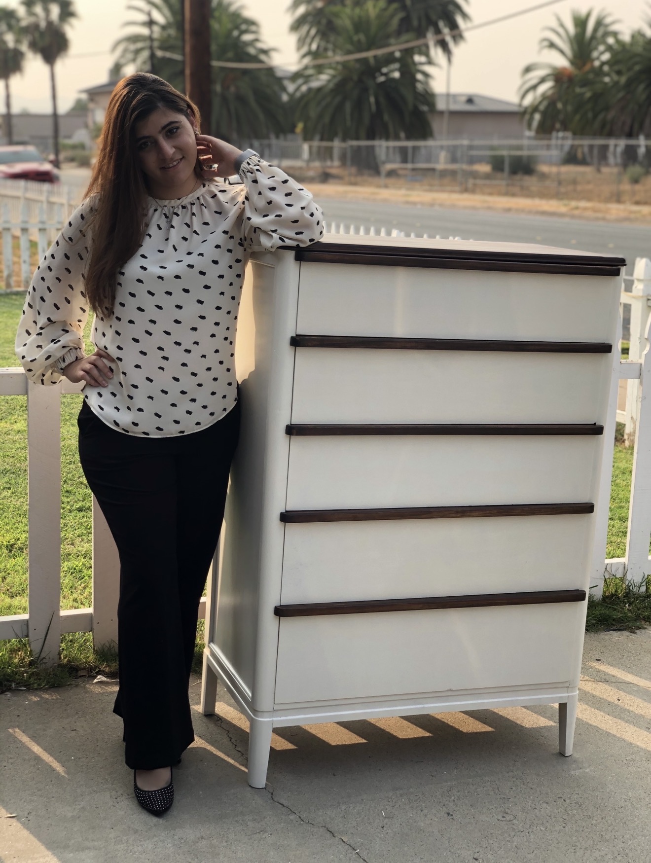Woman with long hair leaning on a white chest of drawers outdoors, wearing a polka dot blouse and black pants.