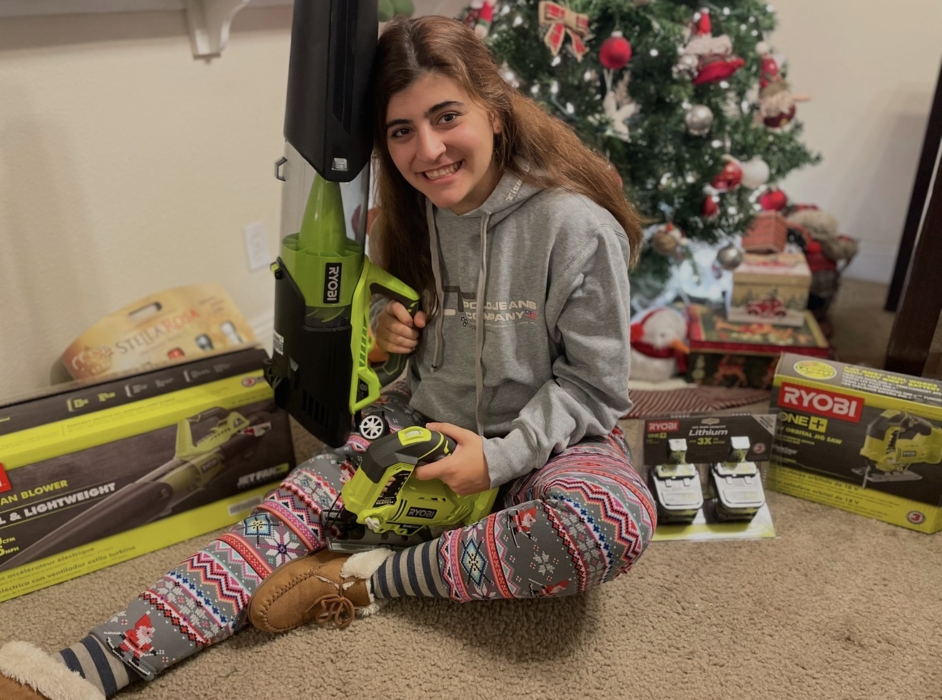 Young girl sitting on carpeted floor, smiling, holding a green and black power tool, with a decorated Christmas tree in background.