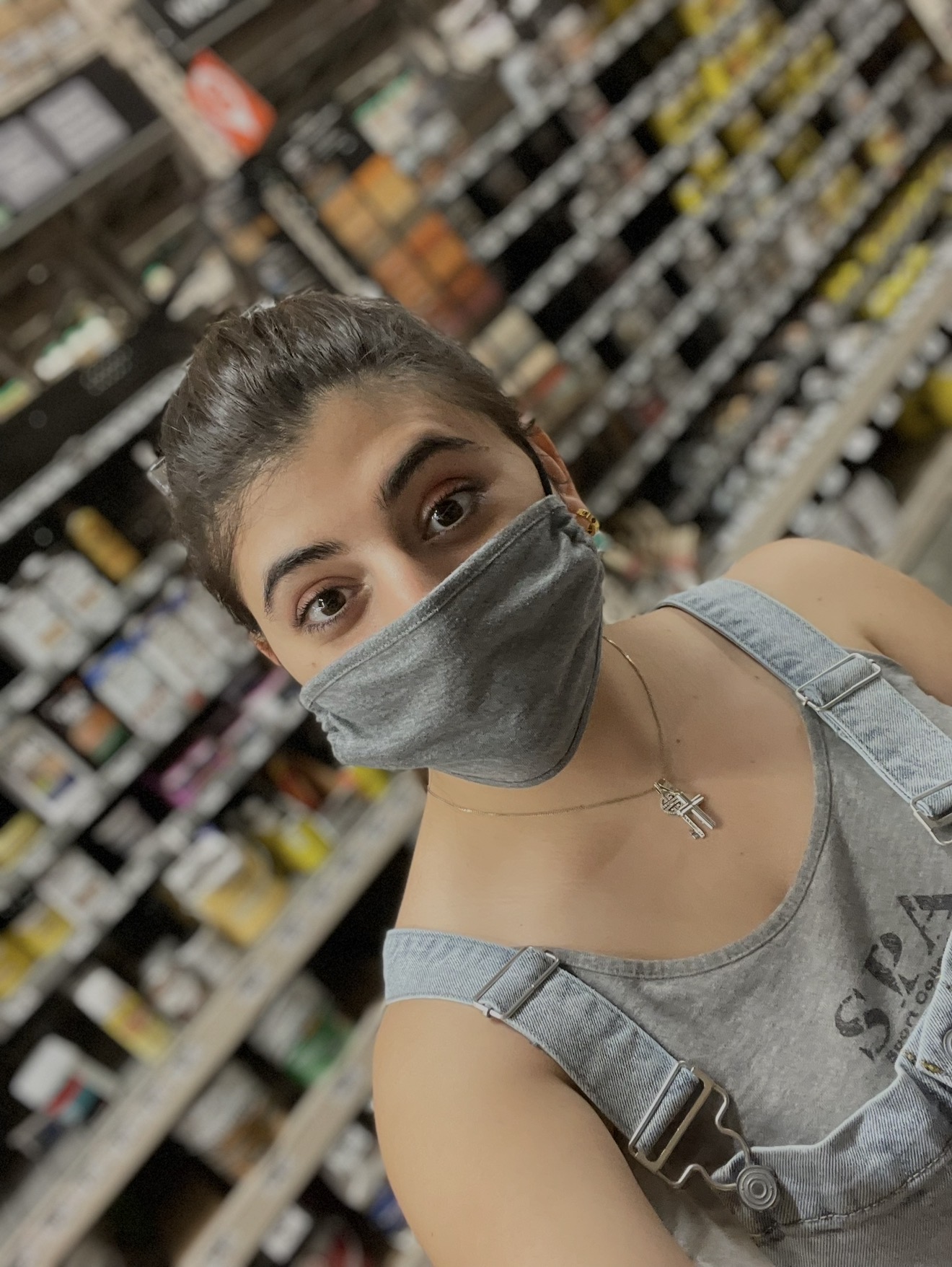 Young woman with dark hair in a bun wearing a gray face mask and denim overalls in a store aisle.