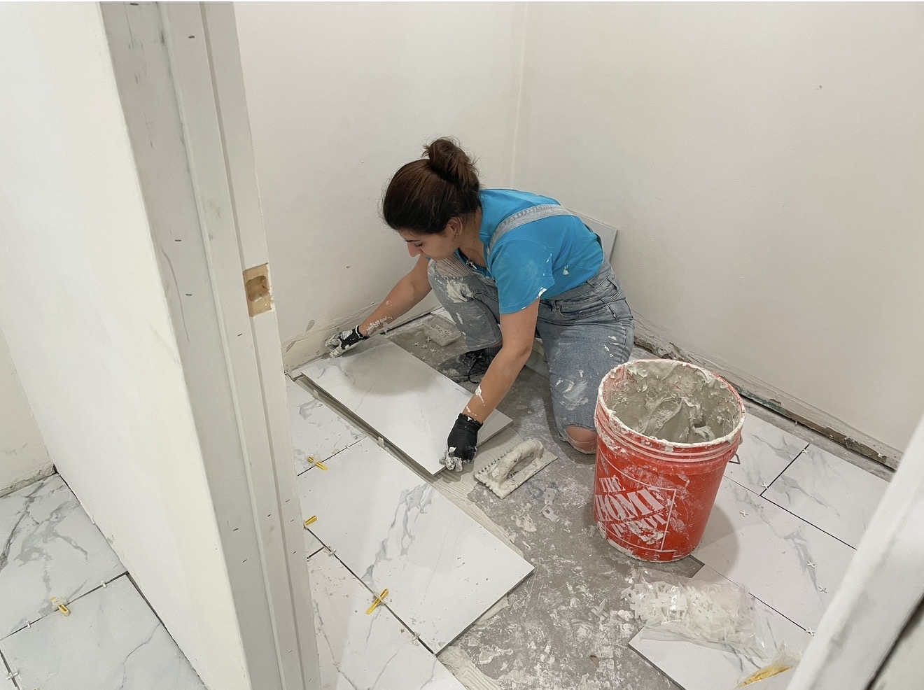 Person kneeling on floor installing tiles, with a bucket nearby, in a room with white walls and marble floor.