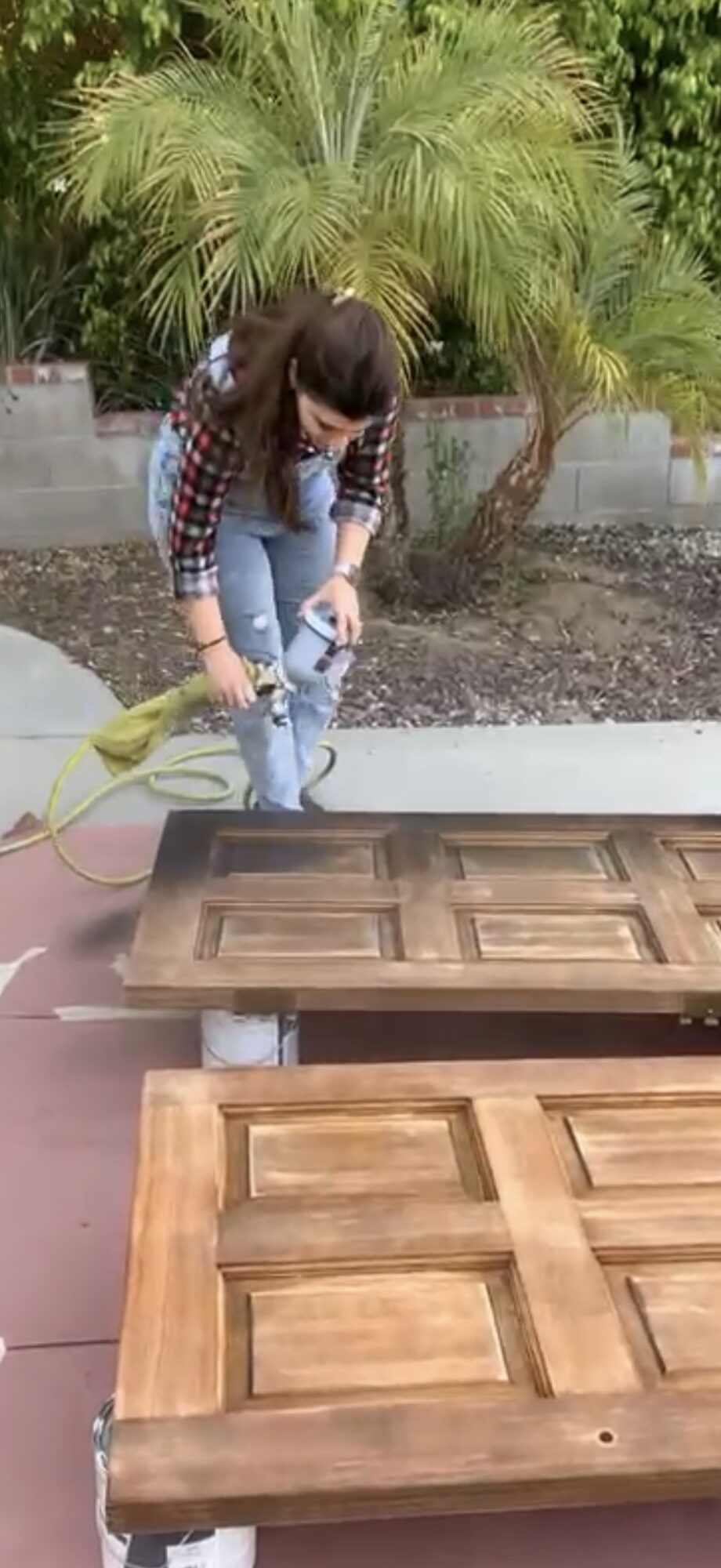 Person spray painting wooden cabinet doors outdoors with plants in background.