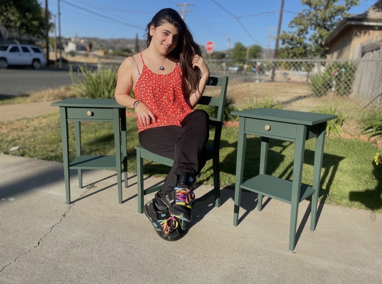 Young woman sitting on a chair outdoors, flanked by two green side tables, with a sunny background and clear sky.