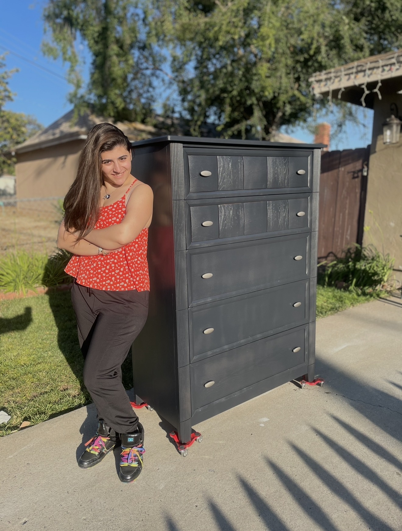 Young woman with long hair in a red polka dot top and black pants stands next to a large black dresser outdoors, casting a shadow.
