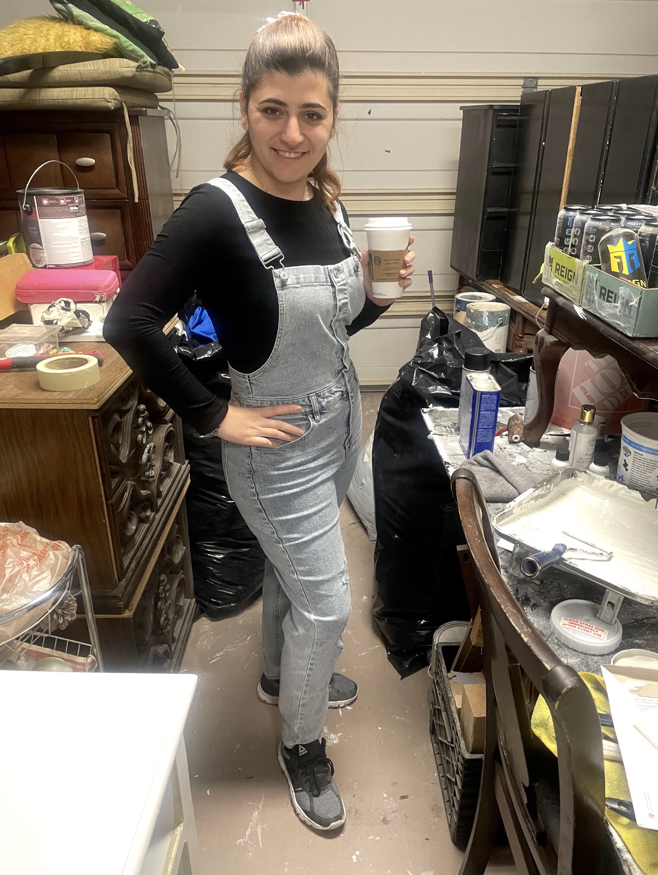 Young woman with a ponytail, smiling, holding a drink, standing in a cluttered workshop or storage room.