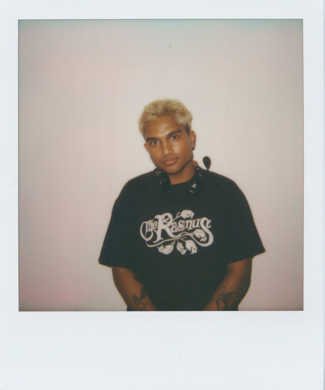 Person with short blonde hair wearing a black T-shirt with white text, standing against a plain white wall.