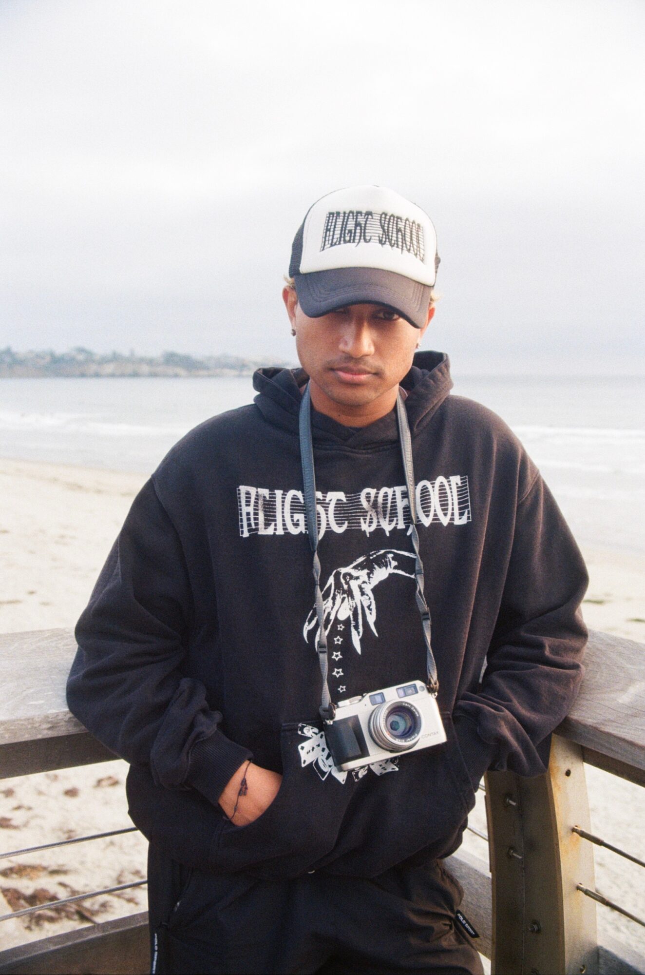 Young man wearing a cap and hoodie, standing outdoors near a beach with water in the background.