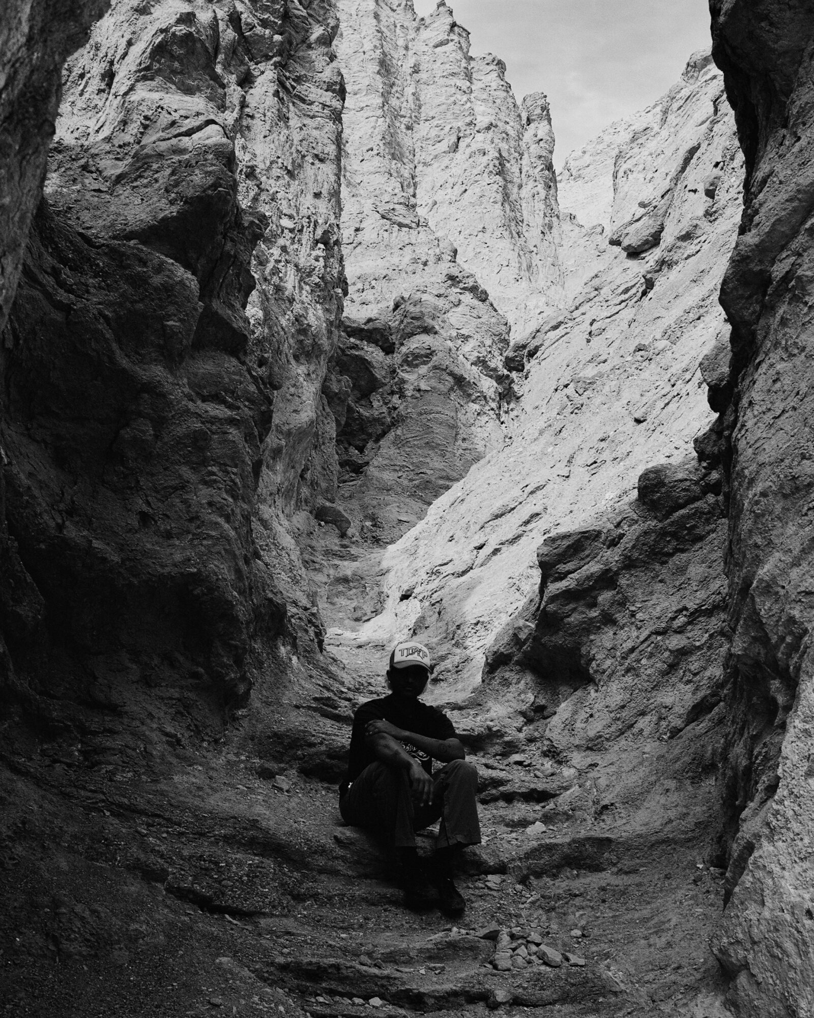 Person sitting in a narrow canyon with tall rocky walls, black and white photo, natural landscape.