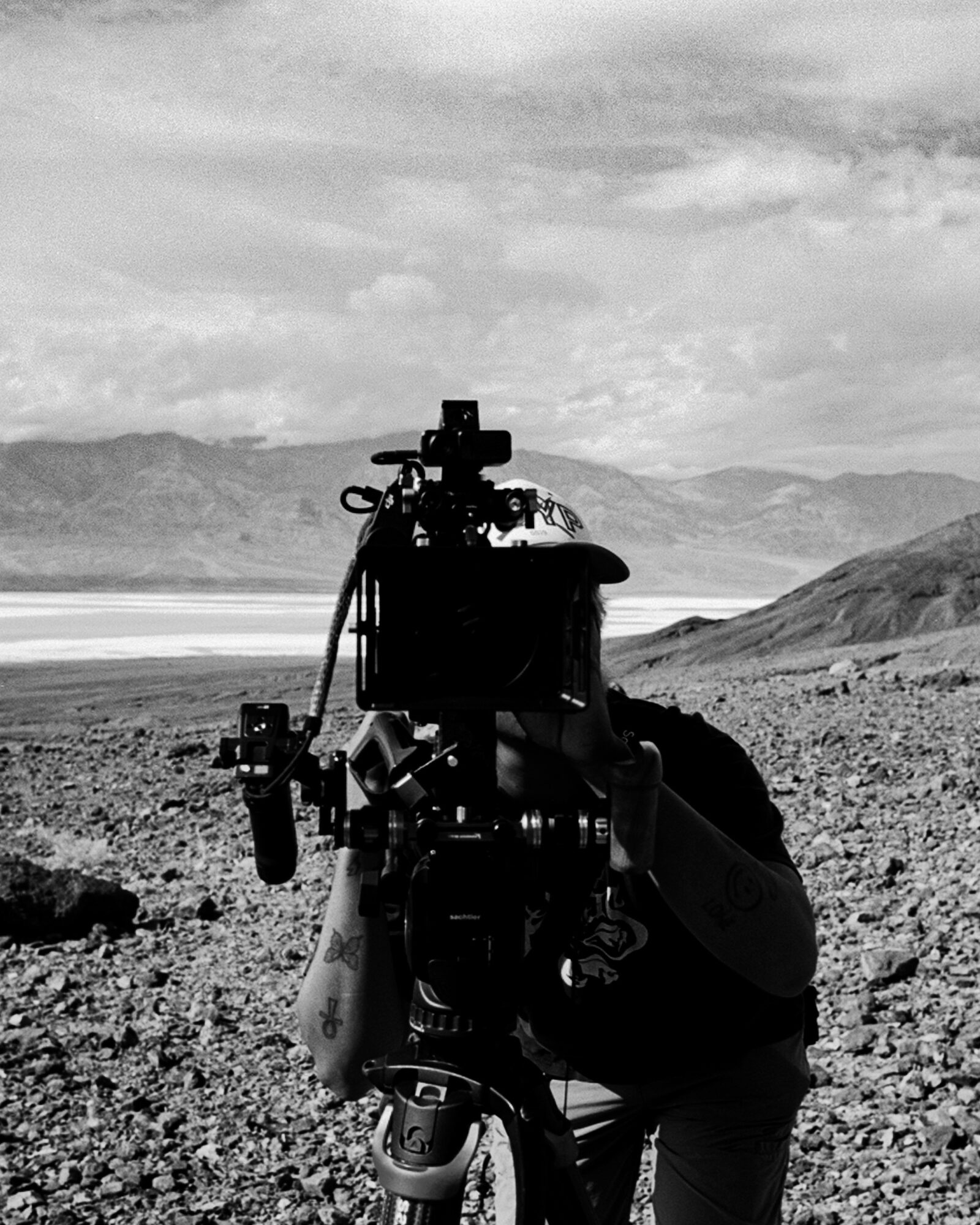 Person operating a camera outdoors with mountains and sky in background, black and white image.