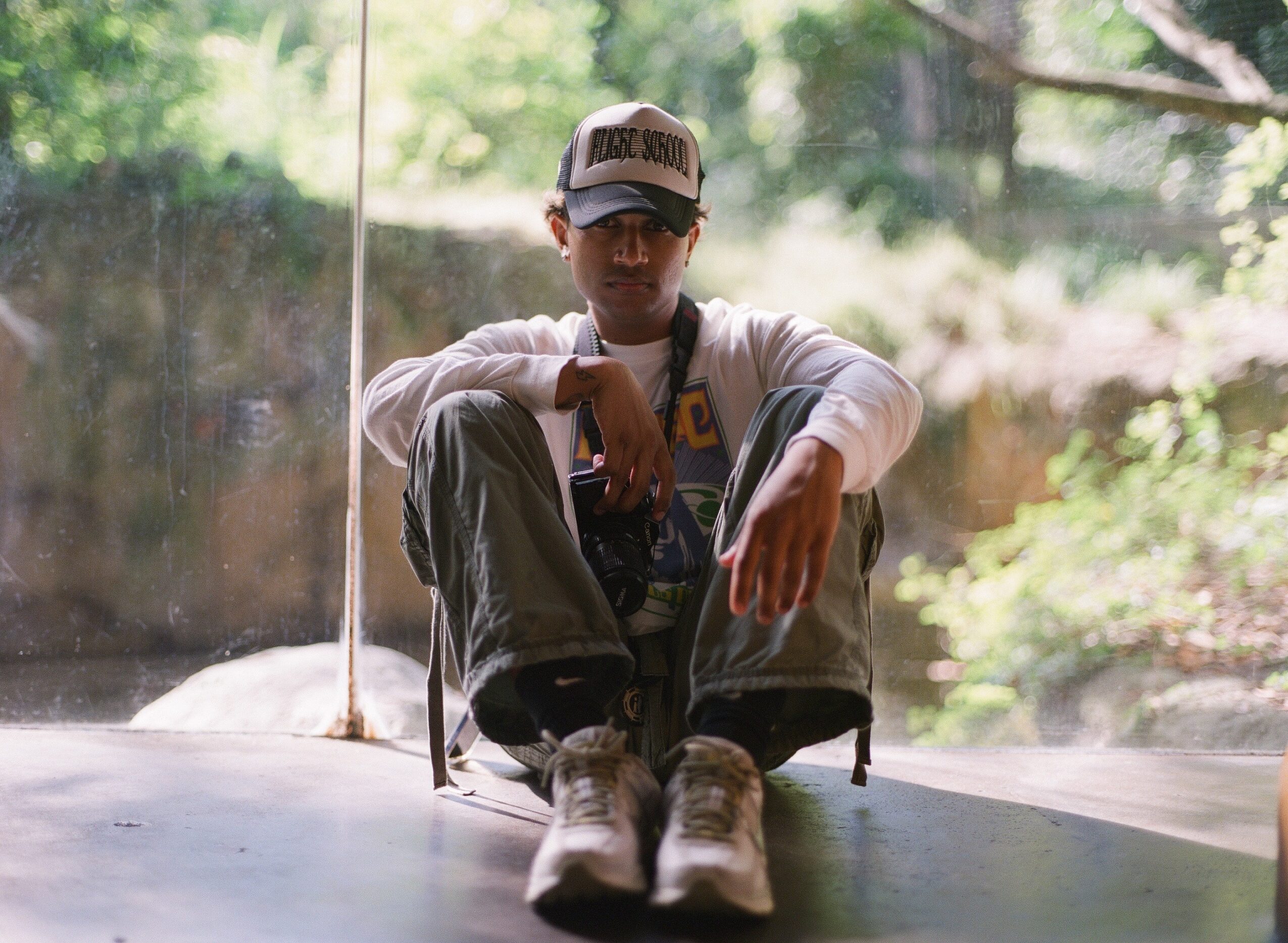 Person sitting with legs crossed, wearing a cap, glasses, and casual clothing, in front of a window with greenery outside.