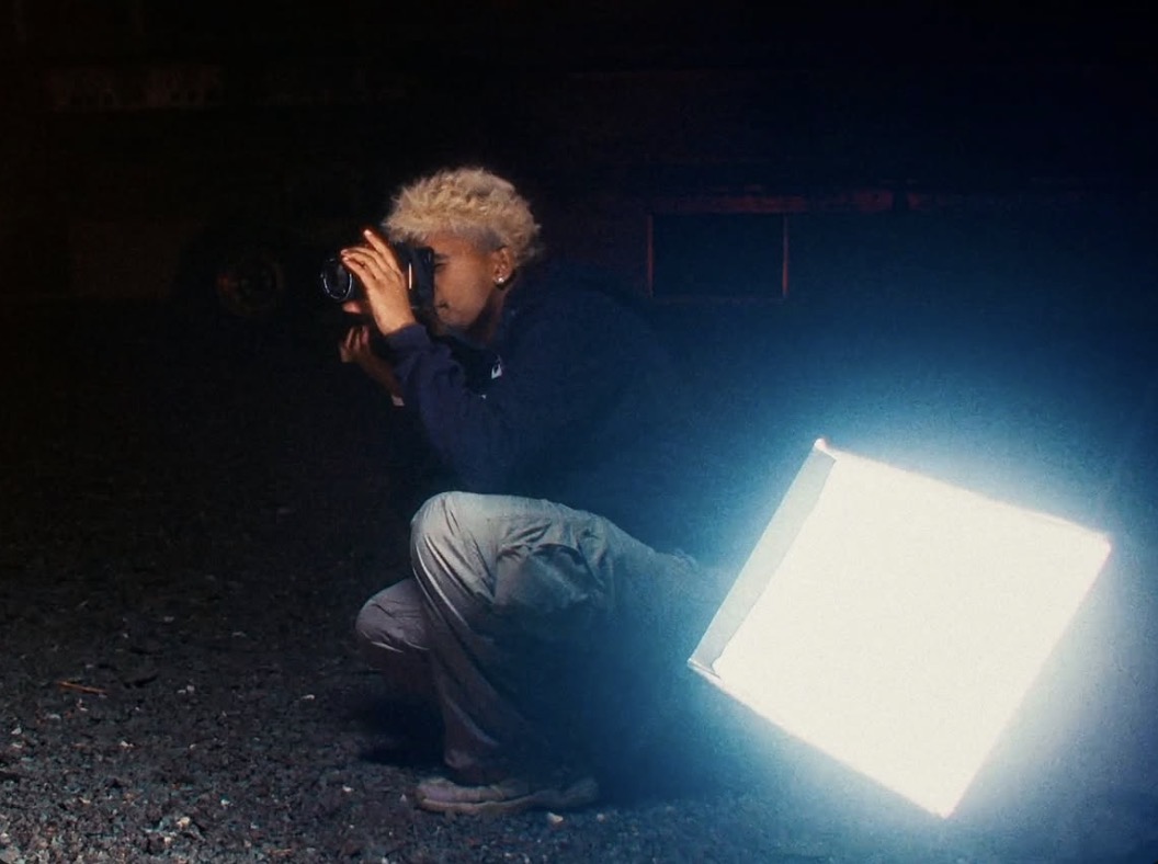Person crouching outdoors at night, holding a camera, illuminated by a bright light source, with dark background.