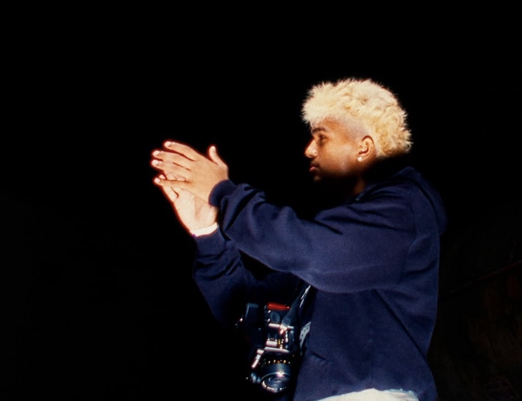 Person with blonde curly hair claps hands against a black background.