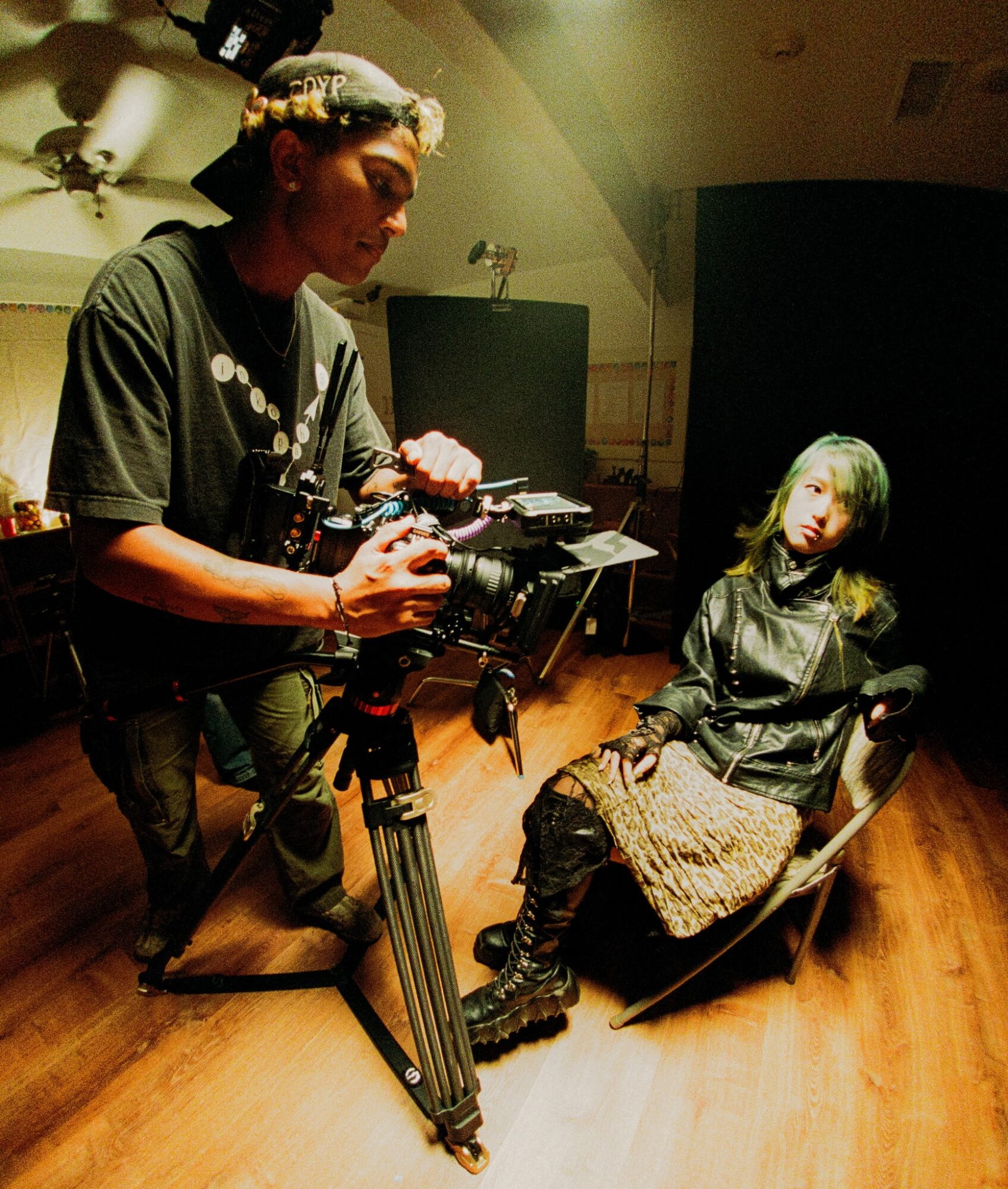 Person operating camera on tripod filming a seated woman with green hair in a room with wooden floor.