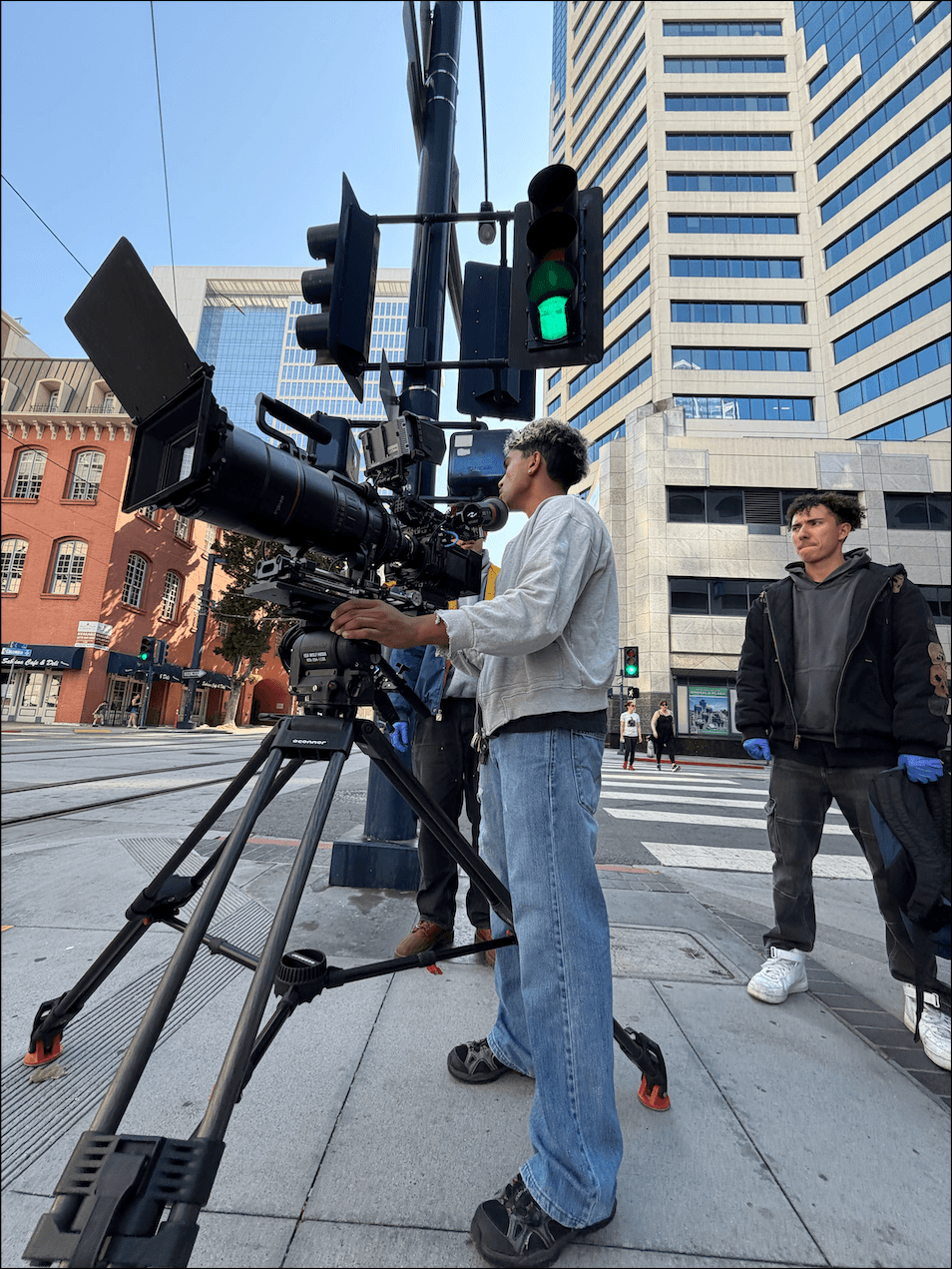 A person operating a professional camera on a city street with tall buildings and pedestrians nearby.