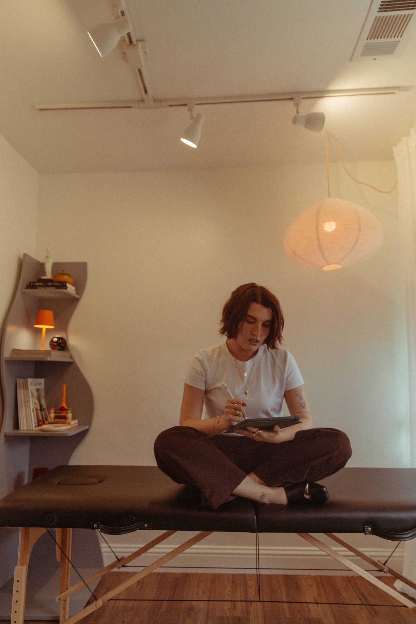 Person sitting cross-legged on a table, reading a book, in a room with a shelf and hanging lamp, white wall background.