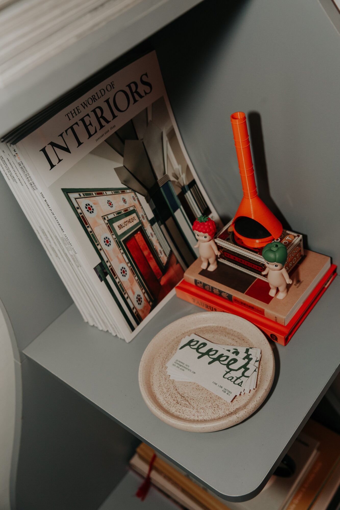 Stack of interior design magazines, a red and black toy, and a small plate with a logo on a gray surface.