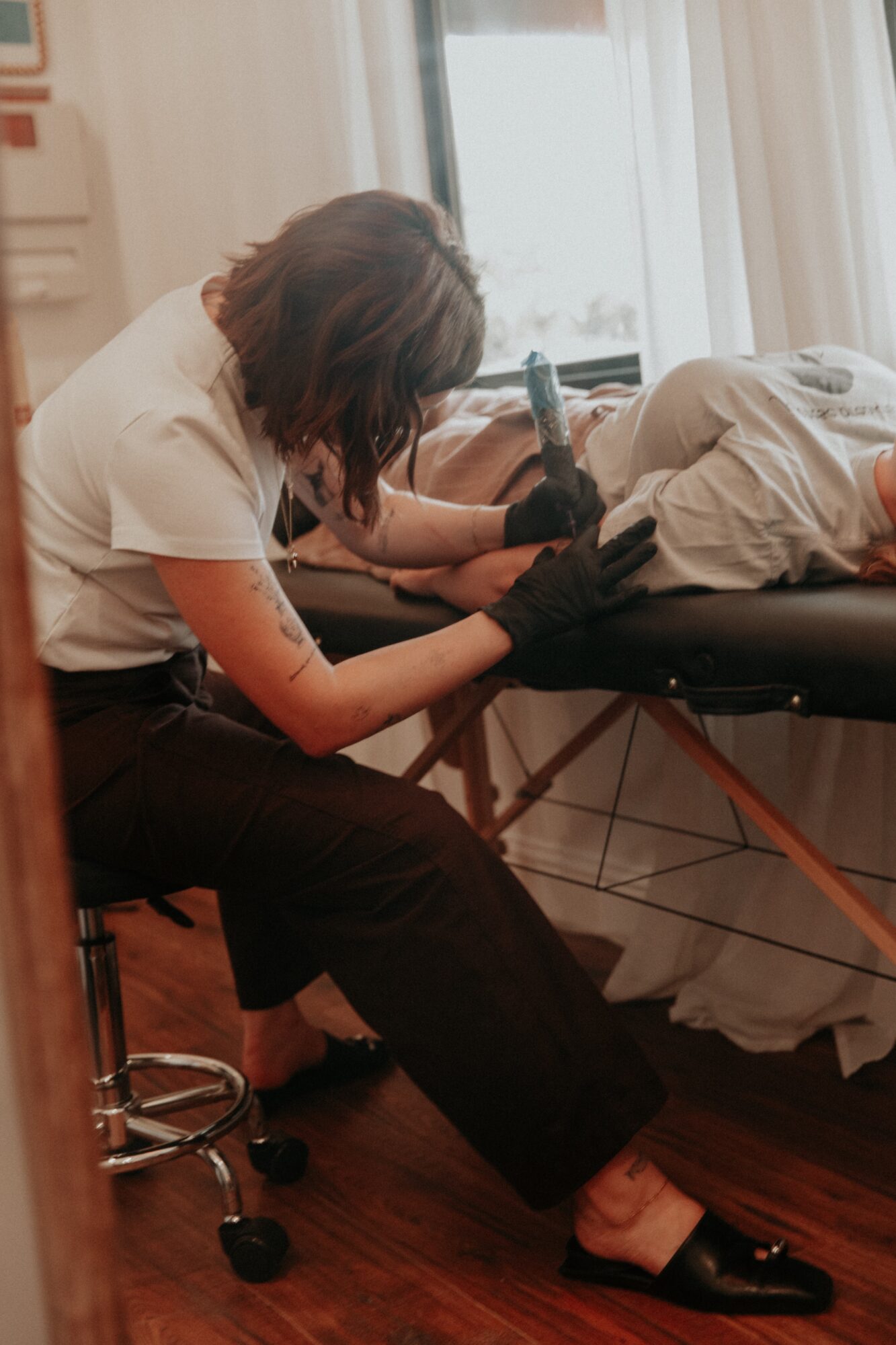 Person with dark hair wearing black gloves tending to a patient lying on a bed, in a room with curtains and wooden floor.