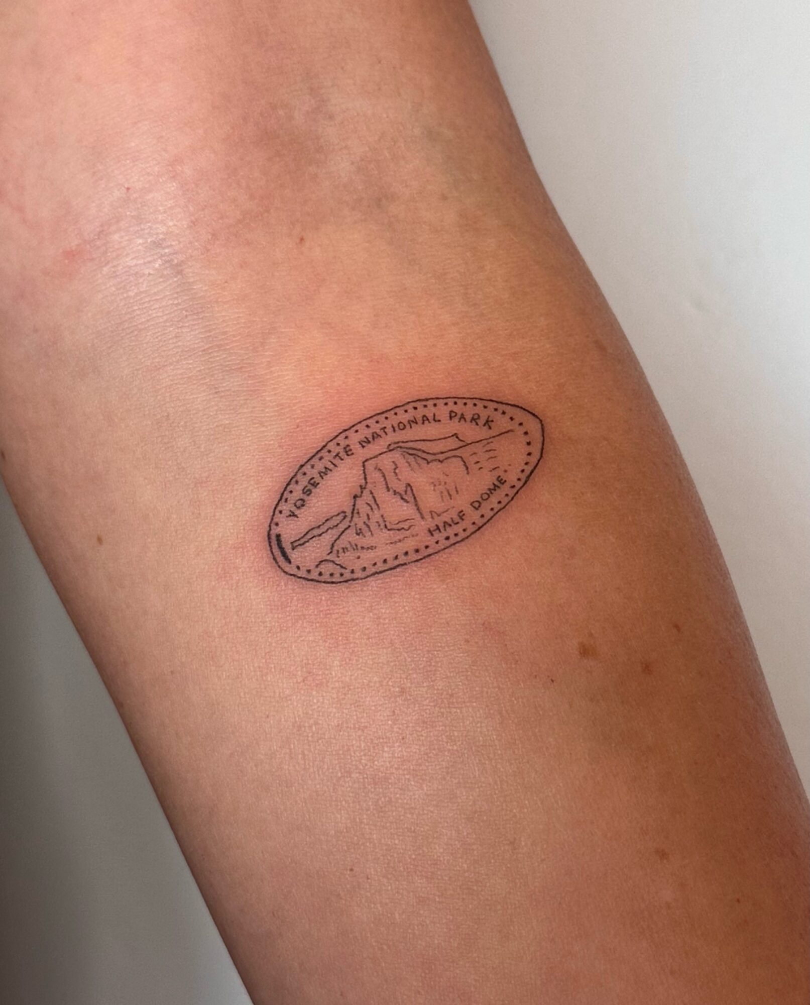 Small oval tattoo of a mountain landscape on skin, with text around the border.