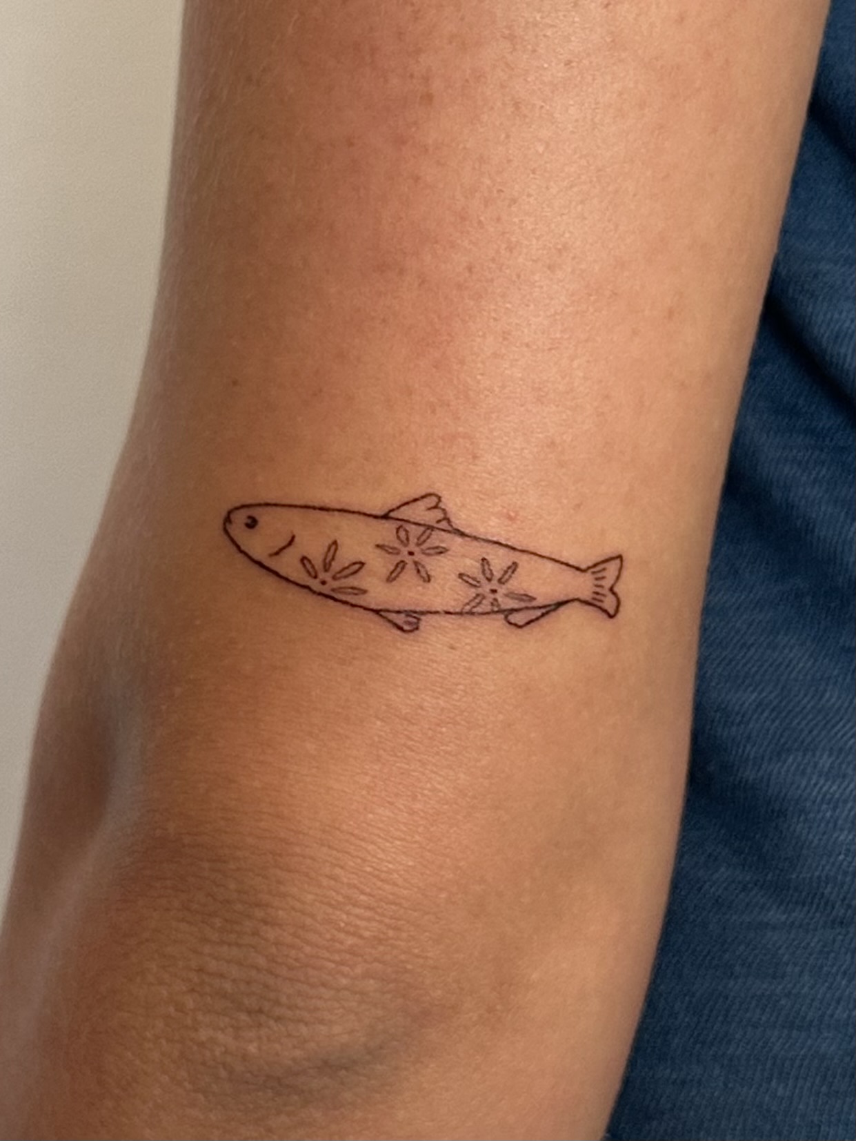 Small fish tattoo with floral details on person's arm, close-up view, skin visible, part of blue clothing seen.
