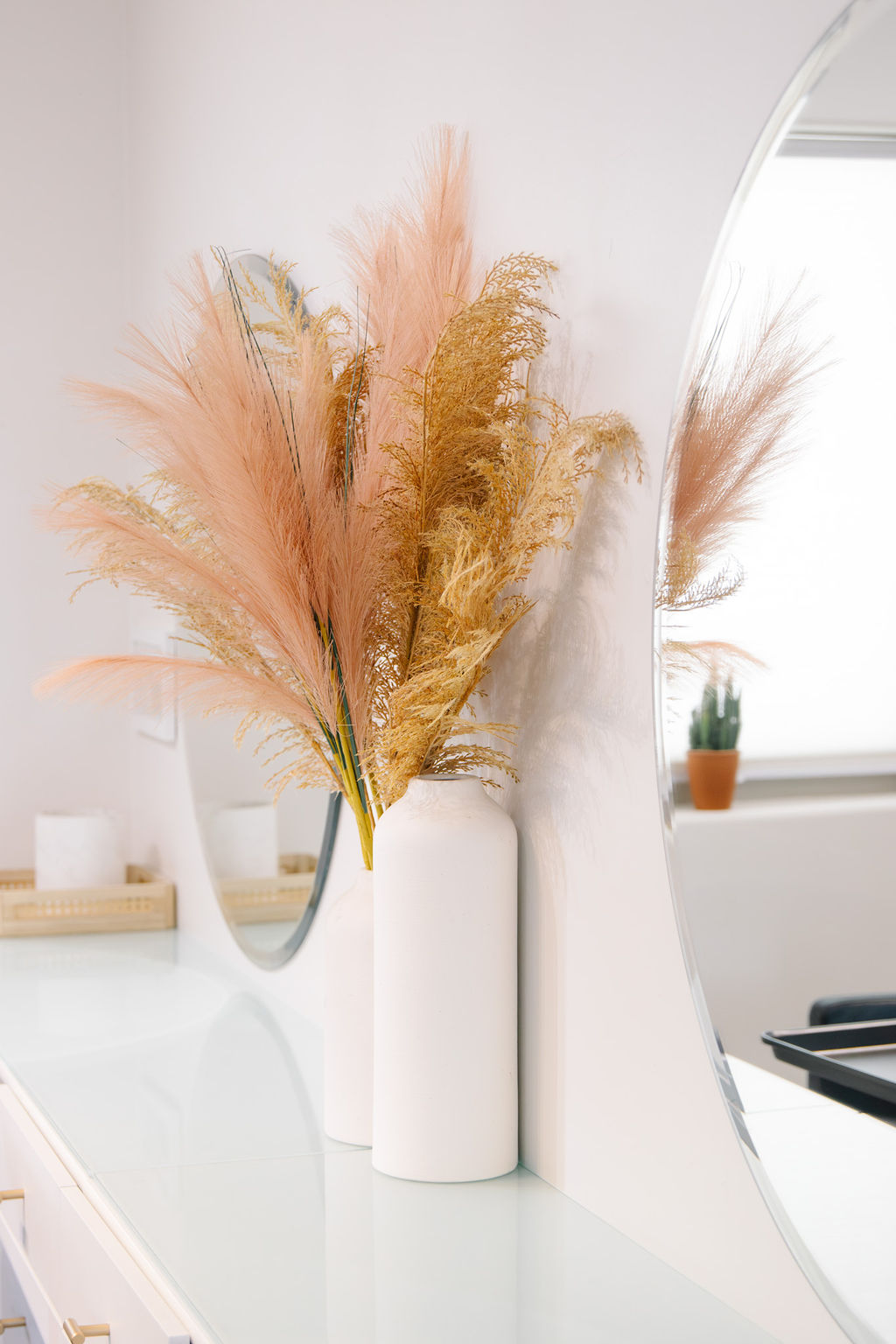 Vase with pink and beige pampas grass on a white surface, mirror, and window in background.