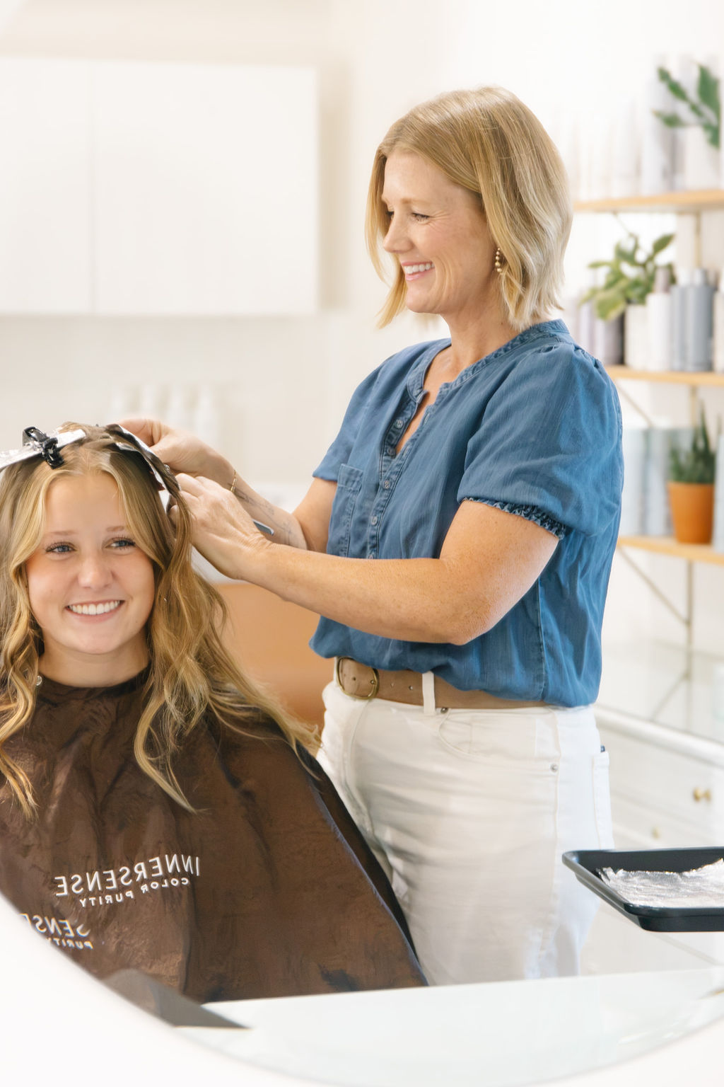 Woman styling a young girl's hair in a bright, modern room with shelves and plants in the background.