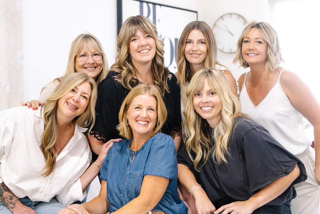 Seven women smiling and posing together indoors, some with arms around each other, in a bright room with a clock and window.