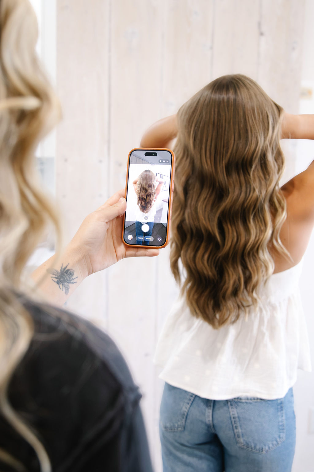Person taking a photo of a girl with long, wavy hair using a smartphone, standing against a light-colored wall.