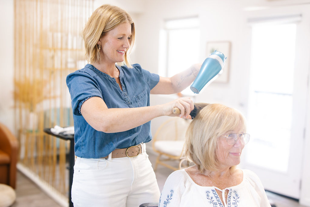 Woman styling an older woman's hair with a spray bottle in a bright room.