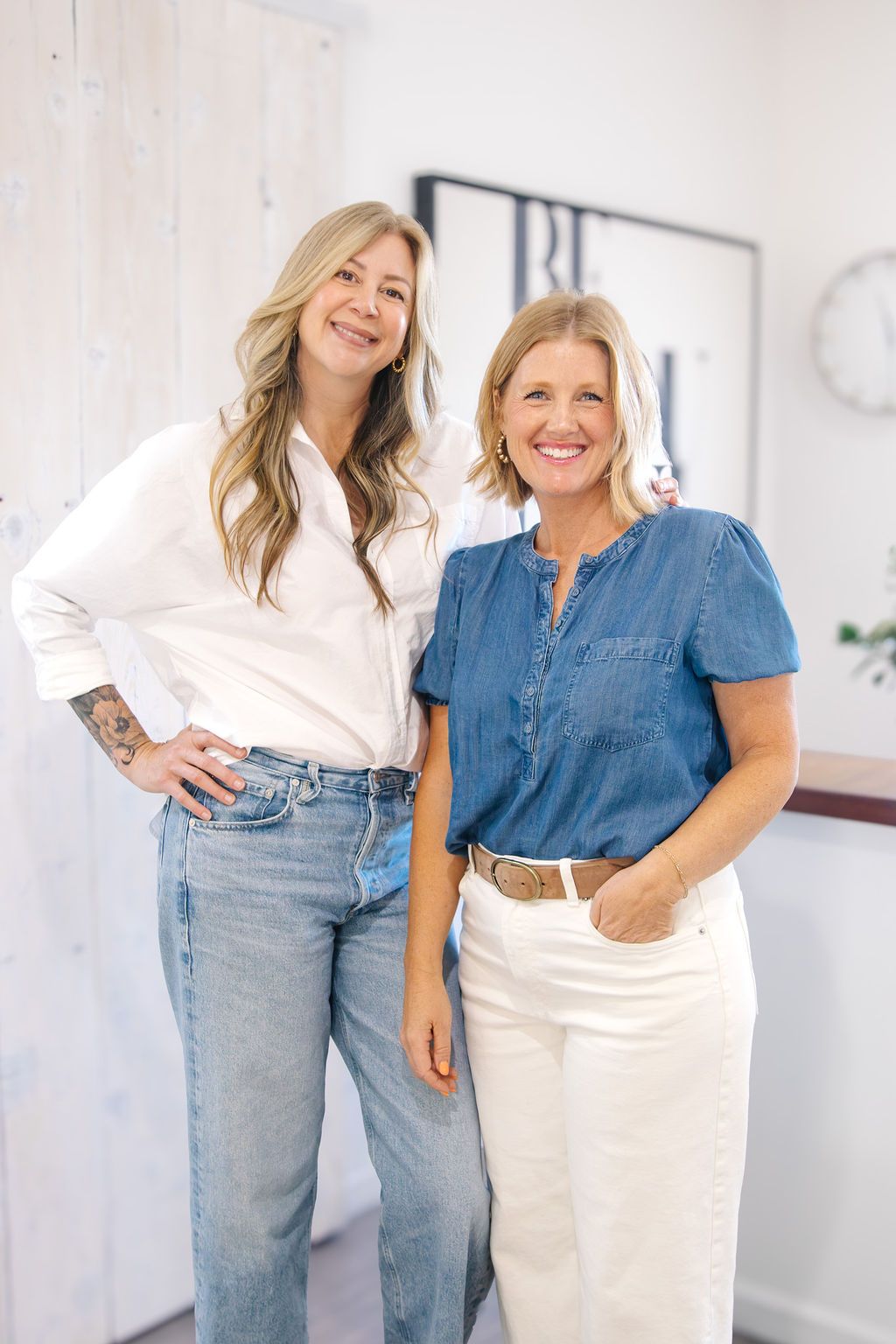Two women smiling indoors, one in a white shirt and jeans, the other in a blue top and white pants.