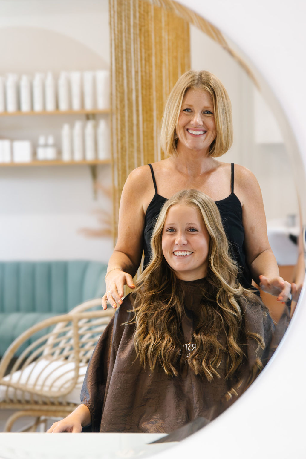 A woman and a girl with long hair smiling in a salon, reflected in a mirror.