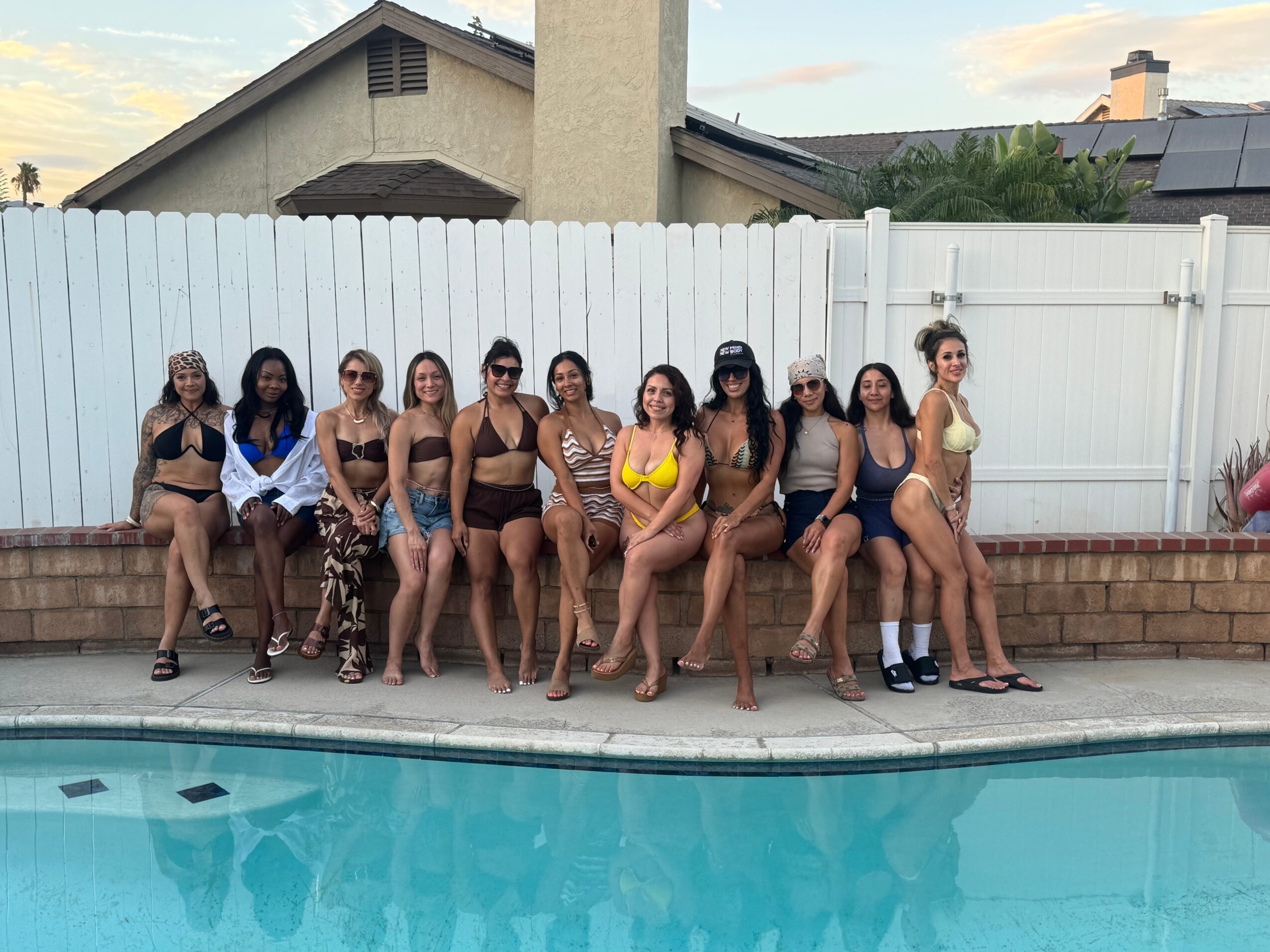 Group of ten women sitting on a brick ledge by a swimming pool, wearing swimsuits and summer clothing, with a white fence behind them.